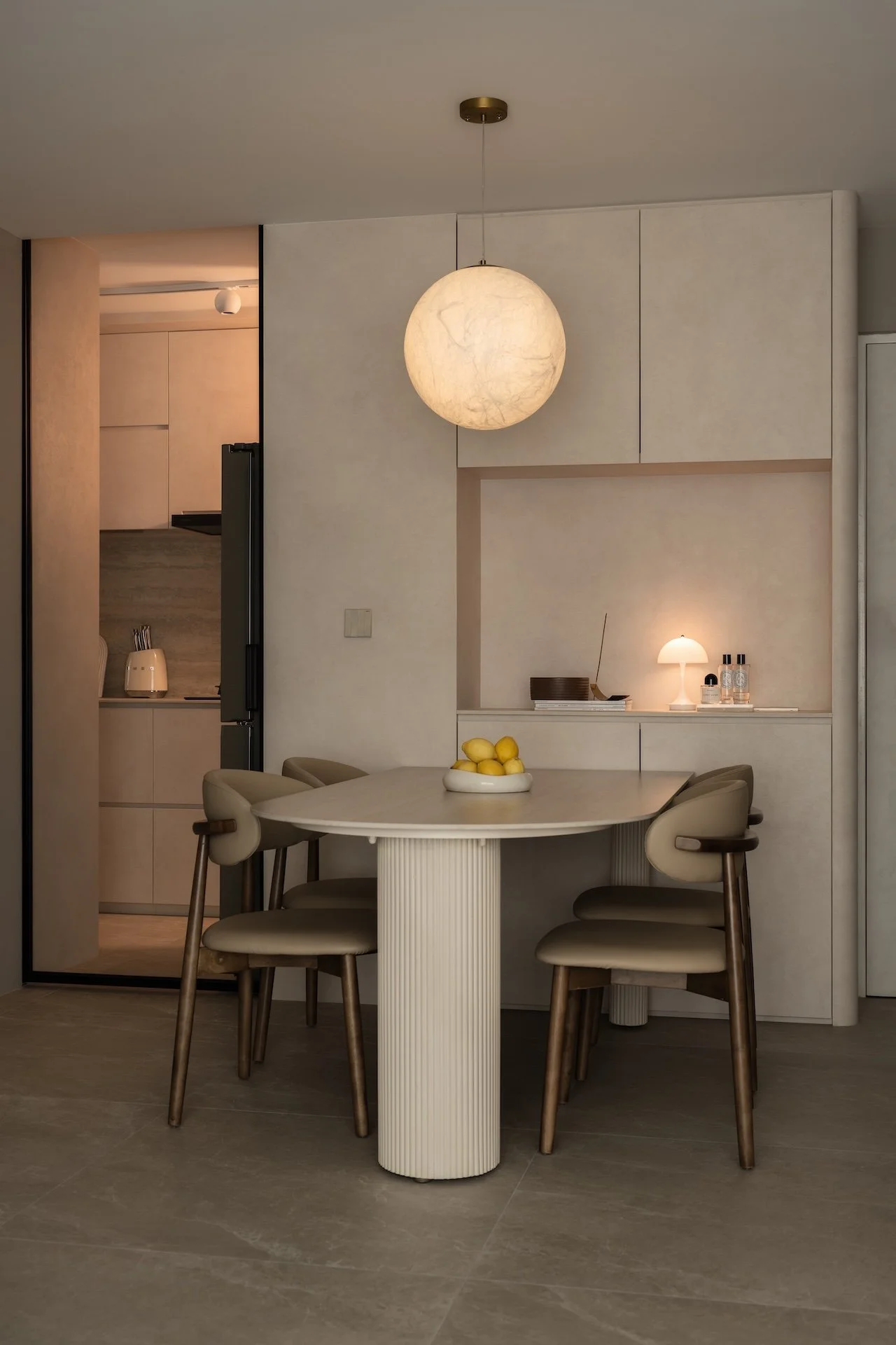 Modern dining area with beige chairs around a rounded table, a bowl of lemons on top, pendant light overhead, and minimal decor including a small table lamp and bottles in a recessed wall nook.