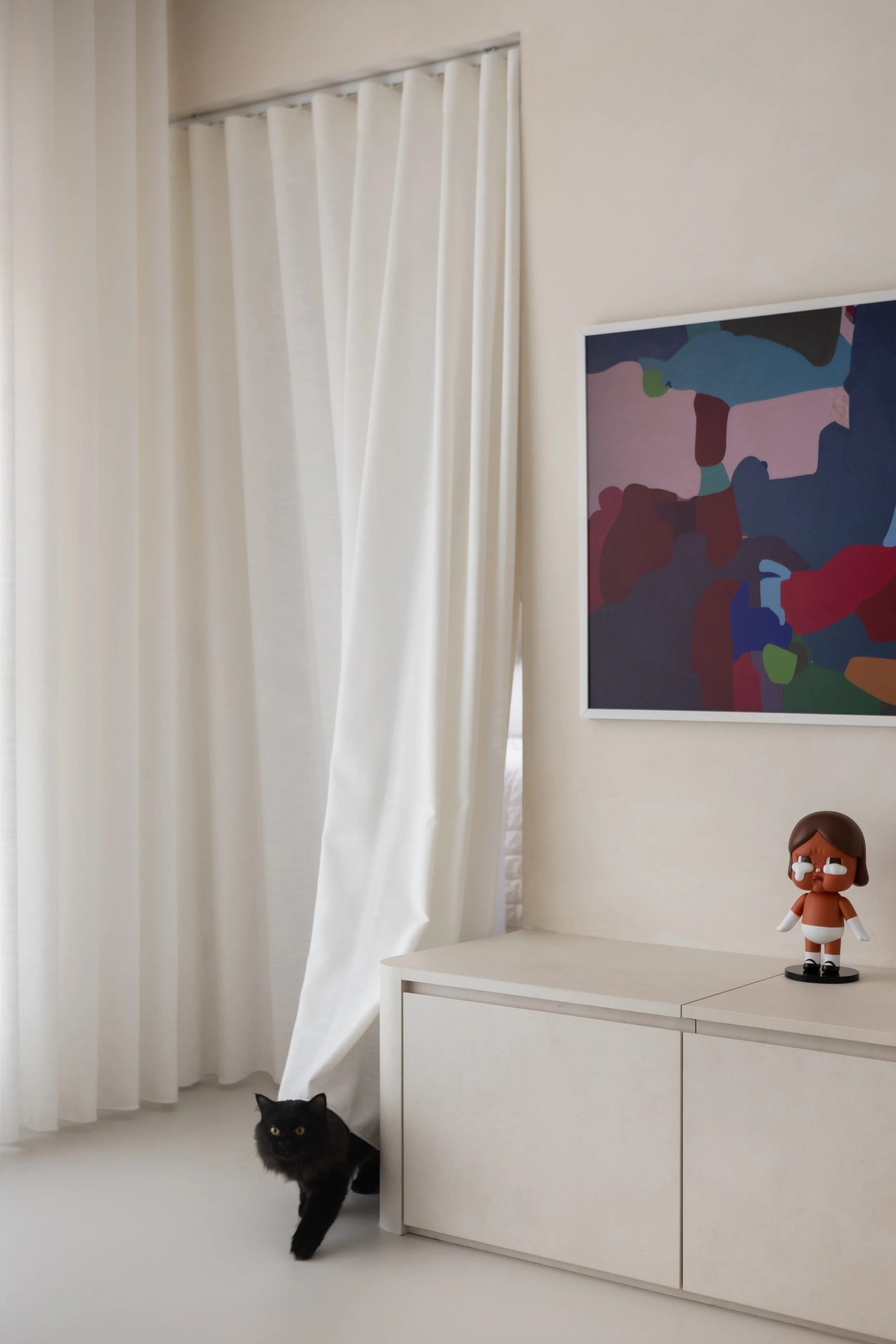 A room with white curtains, abstract colorful painting on the wall, a white cabinet with a small figurine of a girl with glasses, and a black cat walking on the floor.