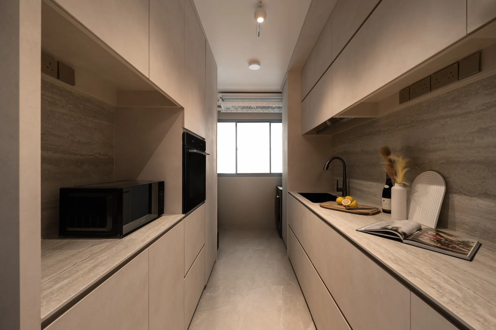 Modern, minimalist kitchen with beige cabinetry, a black sink, and a window at the end. The countertop displays a white vase with dried plants, a bottle, a wooden cutting board with lemons, and an open magazine.