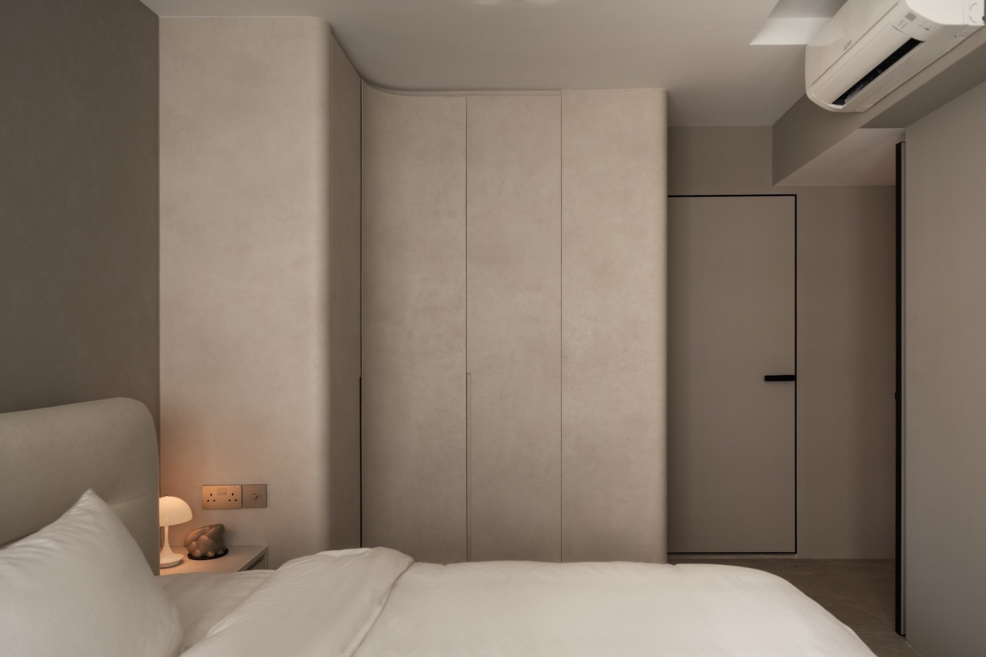 A minimalist bedroom with a white bed, a small bedside lamp, beige walls, a closet with beige doors, and a wall-mounted air conditioning unit.