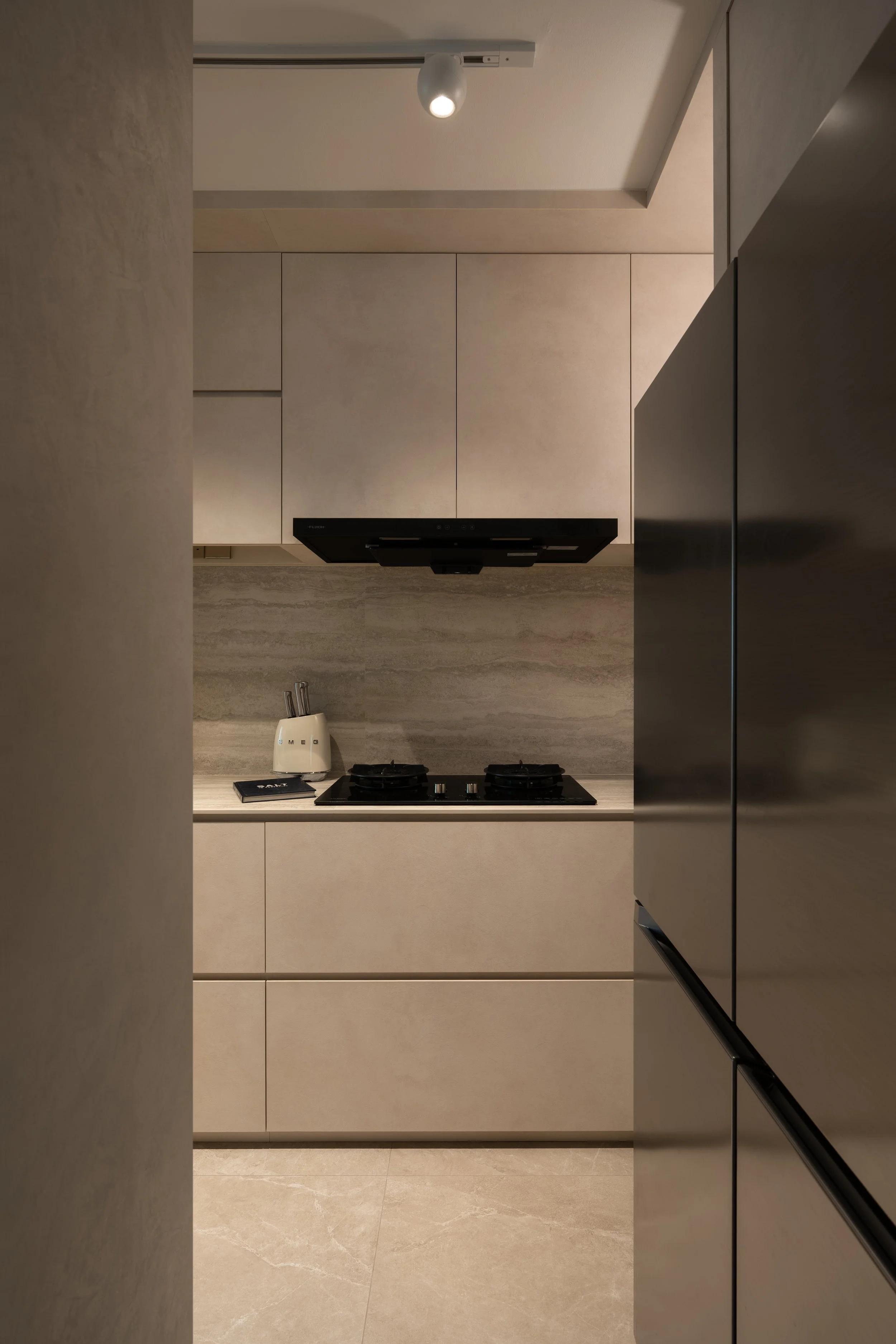 Modern kitchen with beige cabinets, black stovetop, toaster with knives, and gray tiled backsplash, viewed through a narrow opening.