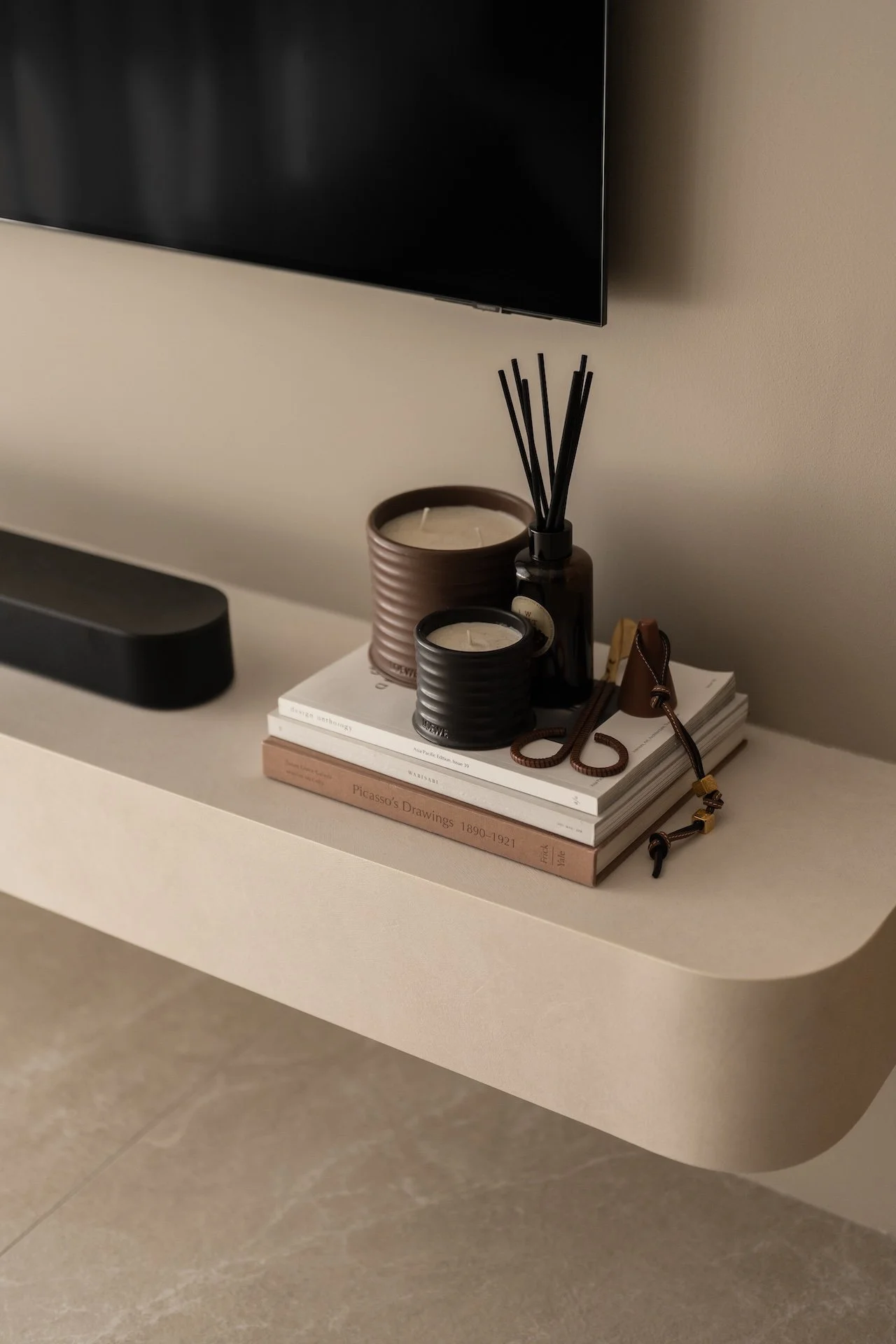 Books, candles, a diffuser with reeds, and a leather strap on a beige floating shelf beneath a wall-mounted TV.