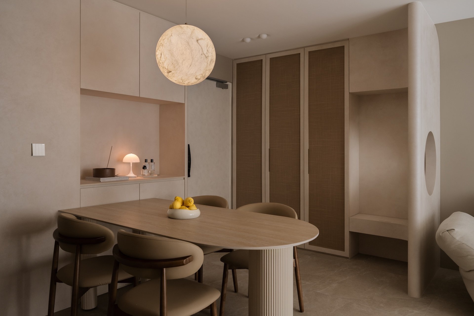 Modern dining room with a wooden table, four beige chairs, a built-in wall shelf with a lamp and decorative items, a large spherical pendant light, and a beige built-in cabinet with sliding doors.
