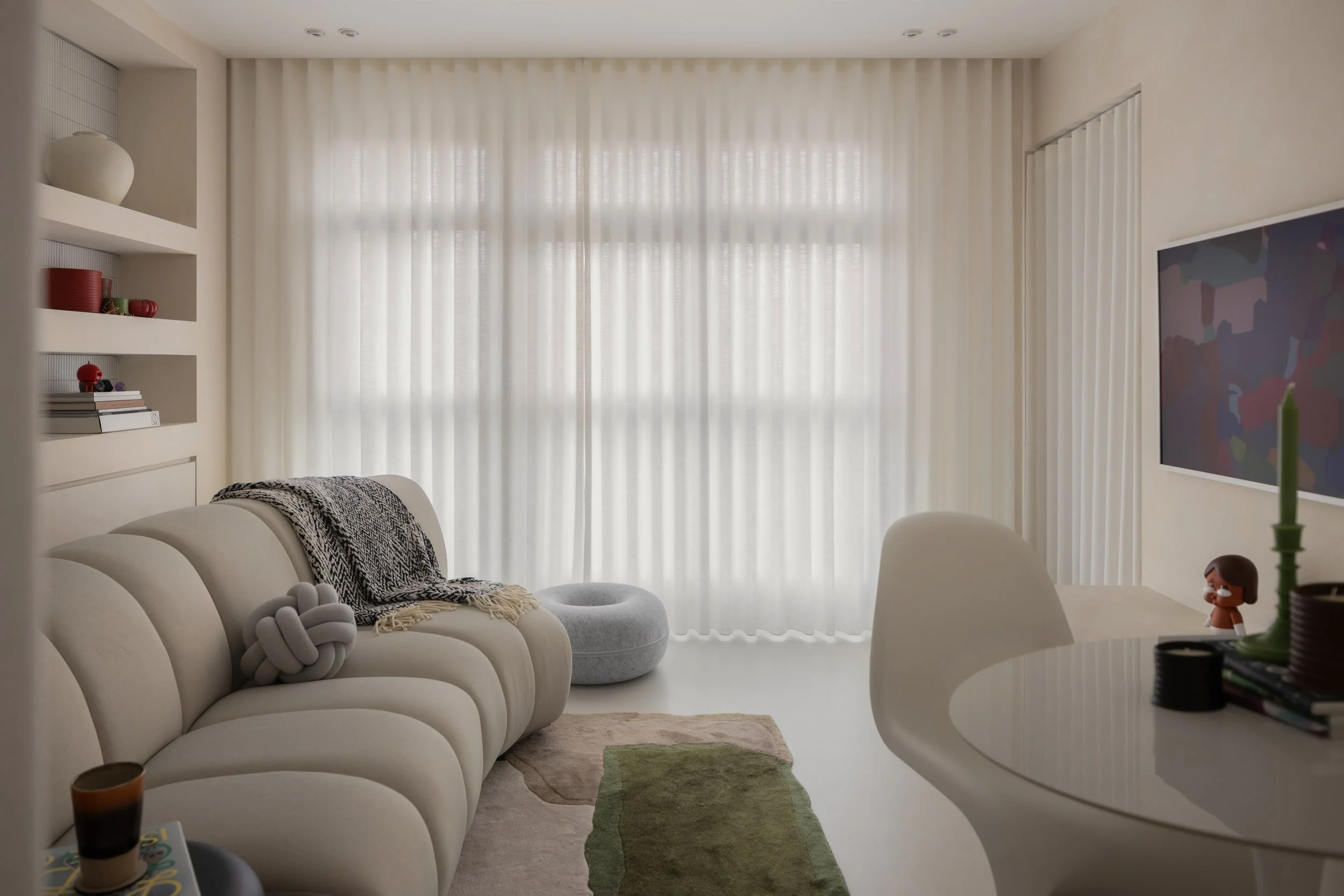 Bright living room with white sofa, grey round ottoman, patterned throw blanket, beige and green area rug, bookshelf with decorative items, large window with sheer curtains, wall-mounted TV, and dining table with white chair and candle