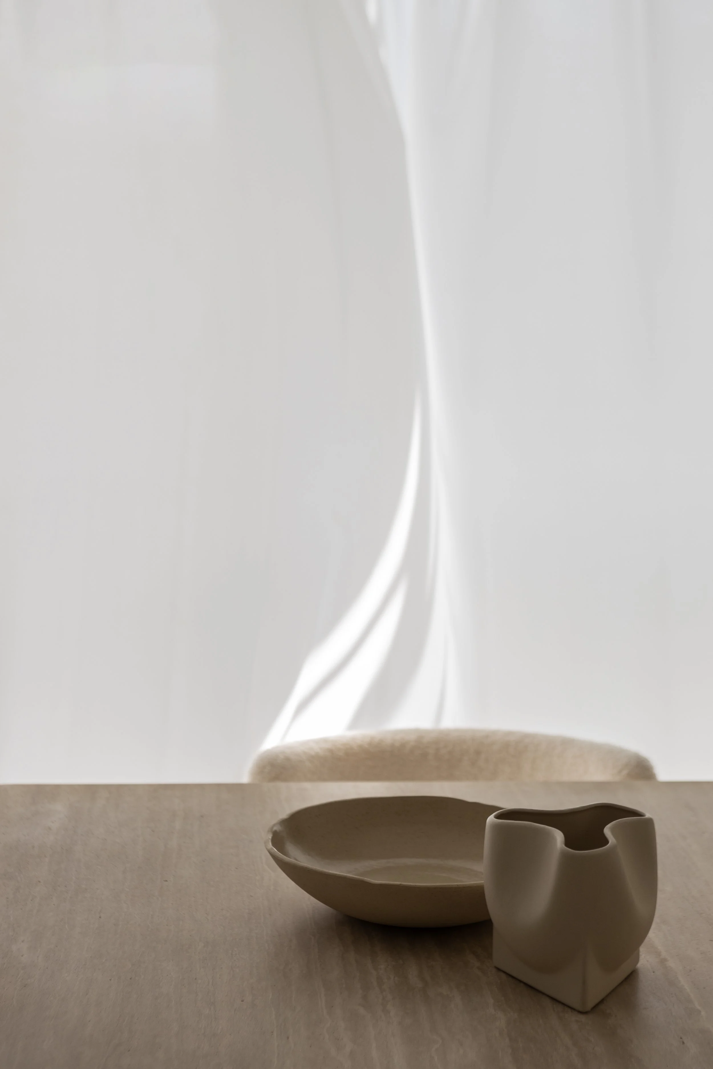A minimalist tabletop scene with a beige shallow bowl and an abstract white ceramic vessel, set against a neutral background with soft lighting.