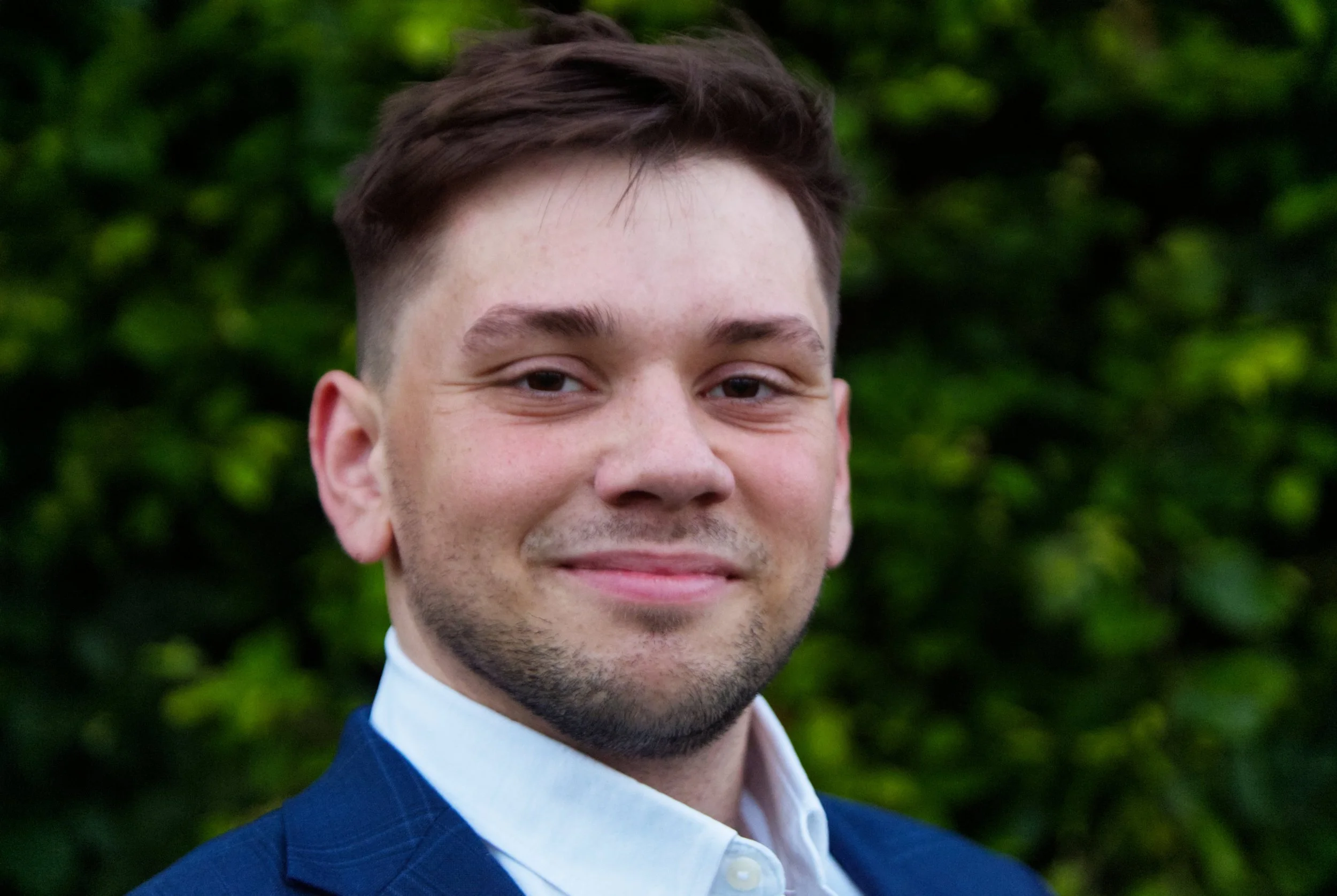 A young man with short brown hair, a light beard, and light skin, wearing a blue suit and white shirt, smiling outdoors with blurred green foliage in the background.
