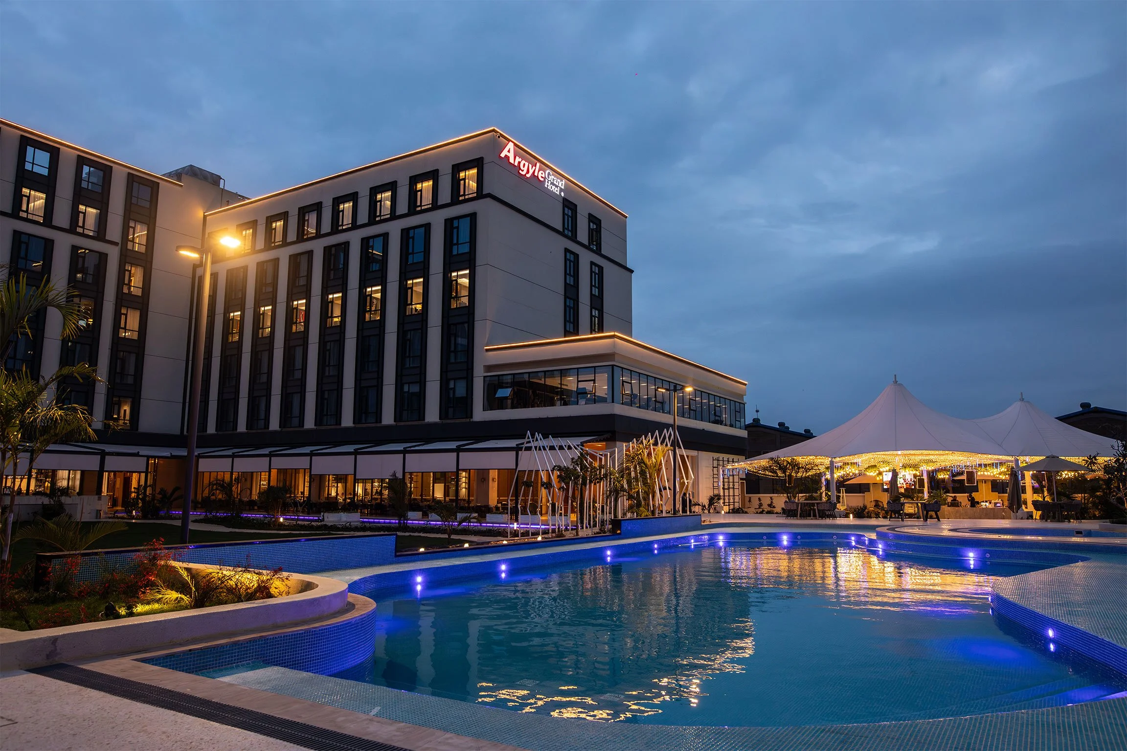 Nighttime view of the Argyle Grand Hotel with a lit swimming pool and a white pavilion tent outside, illuminated with blue and yellow lights, under a cloudy sky.