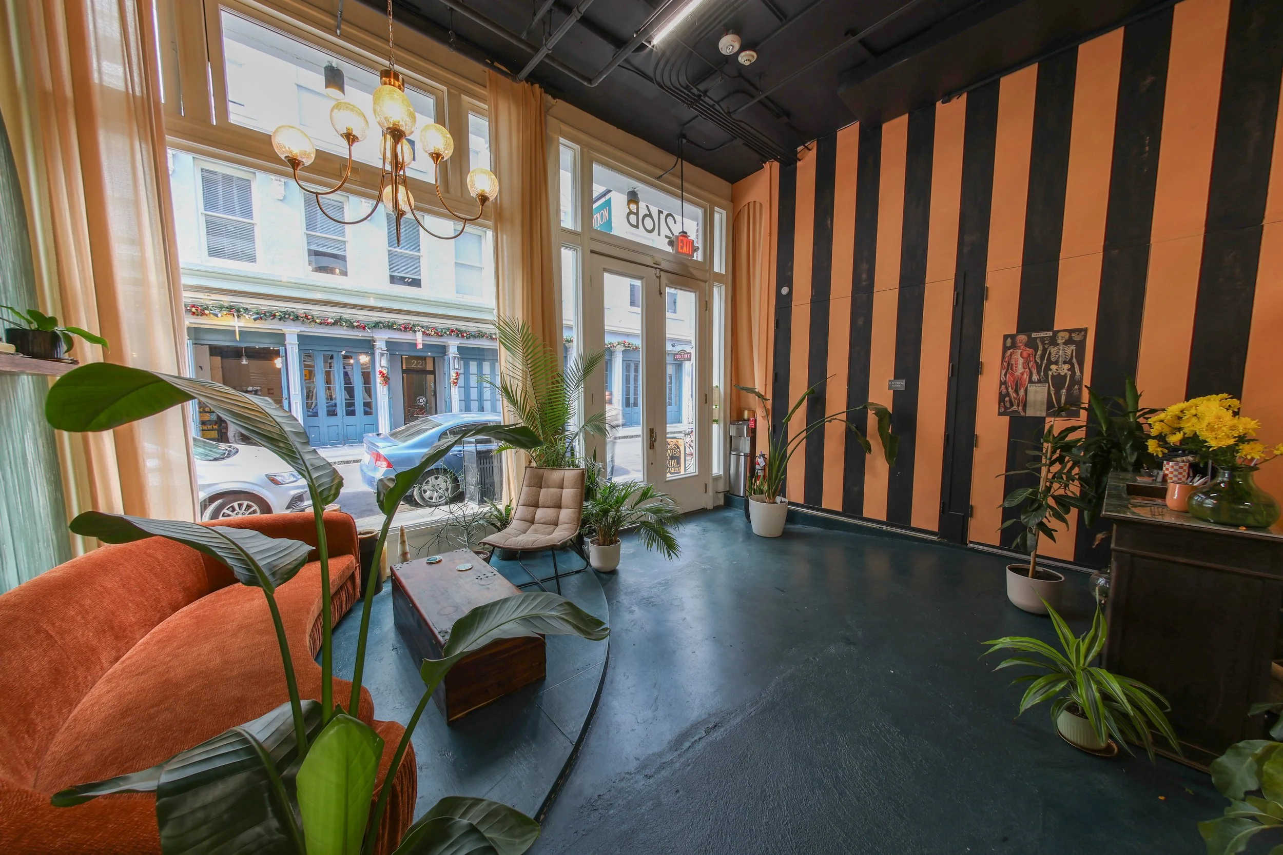 Interior view of a cozy cafe with large windows, beige curtains, a black and orange striped accent wall, a chandelier, potted plants, and seating including an orange sofa and a beige chair.