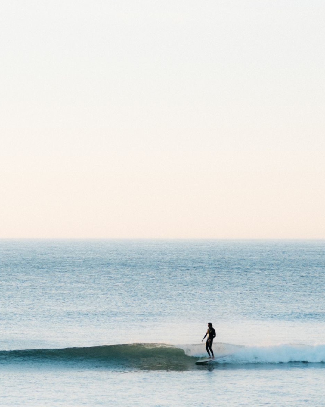 A person surfing on a small wave in the ocean, with a calm sea and clear sky in the background.