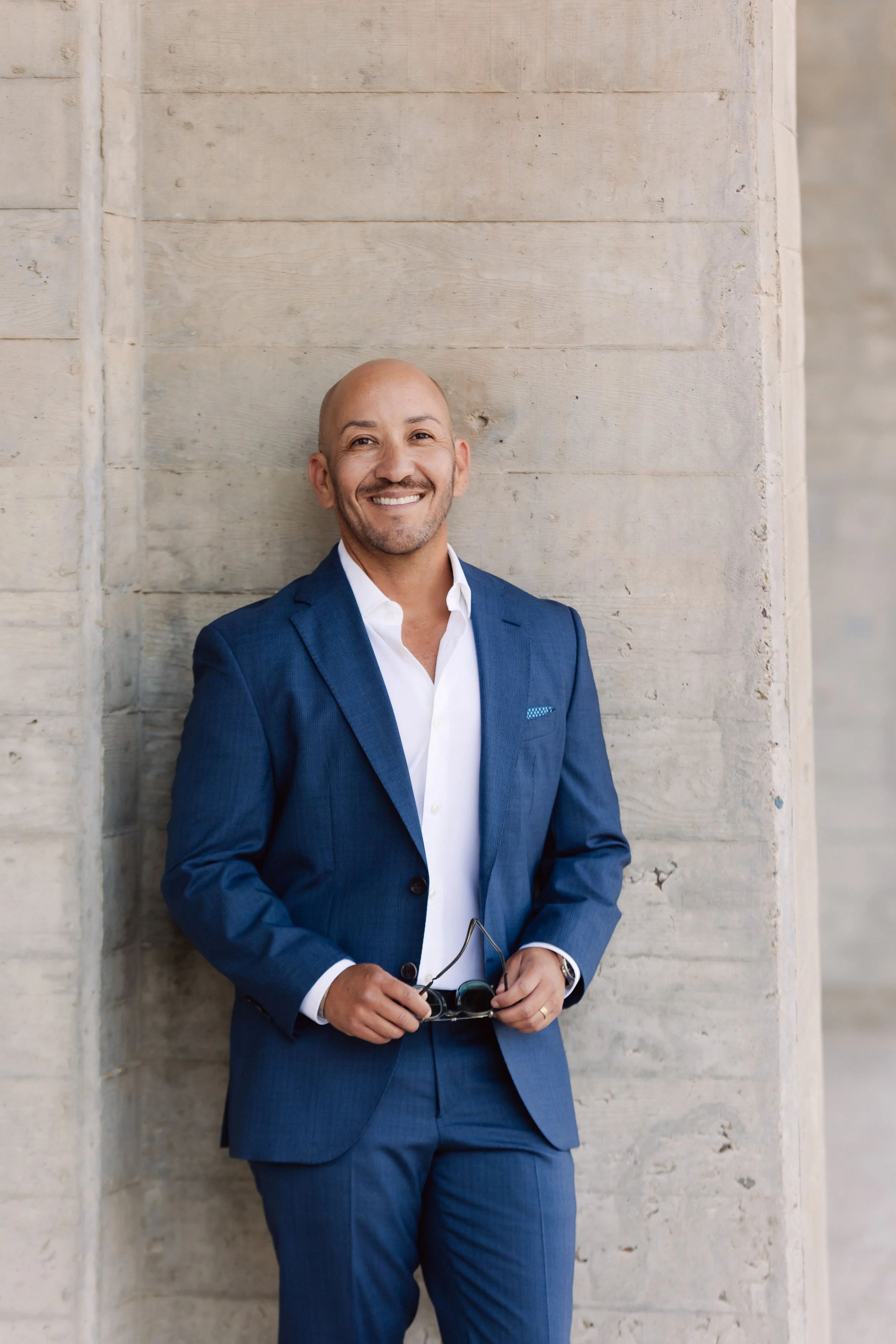 A smiling man in a blue suit and white shirt standing against a concrete wall, holding sunglasses.