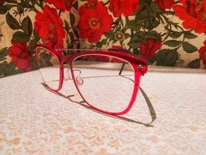 Red eyeglasses with thin frame resting on a surface with a floral red and green patterned background.