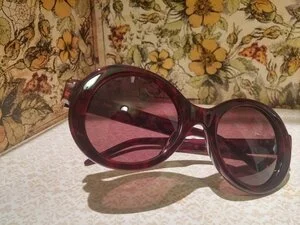 A pair of large, dark purple sunglasses with round lenses resting on a white surface against a floral patterned background.