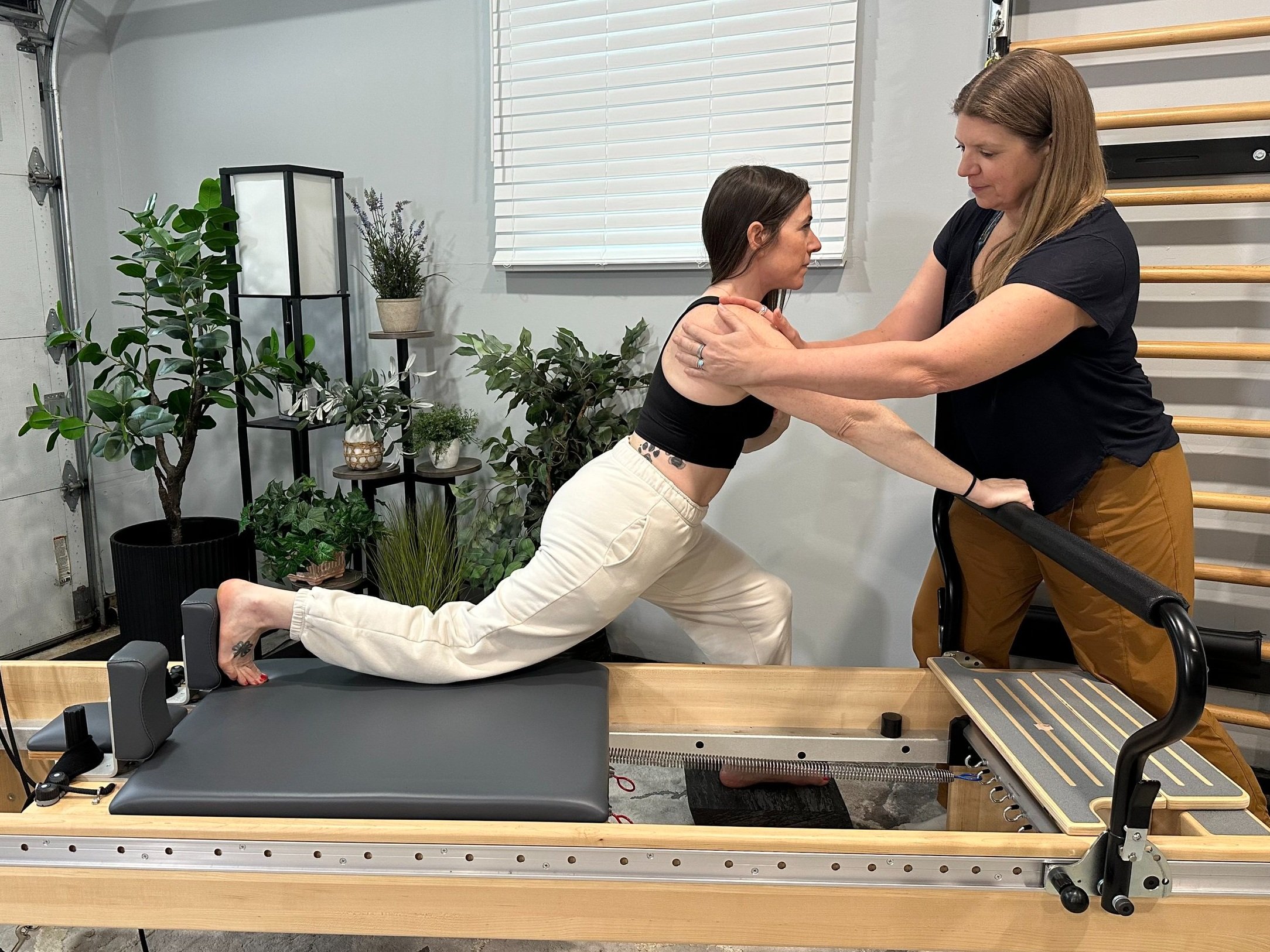 A woman is performing a physical therapy exercise on a reformer machine, supported by a therapist holding her shoulders, in a rehab or fitness studio with plants and exercise equipment.