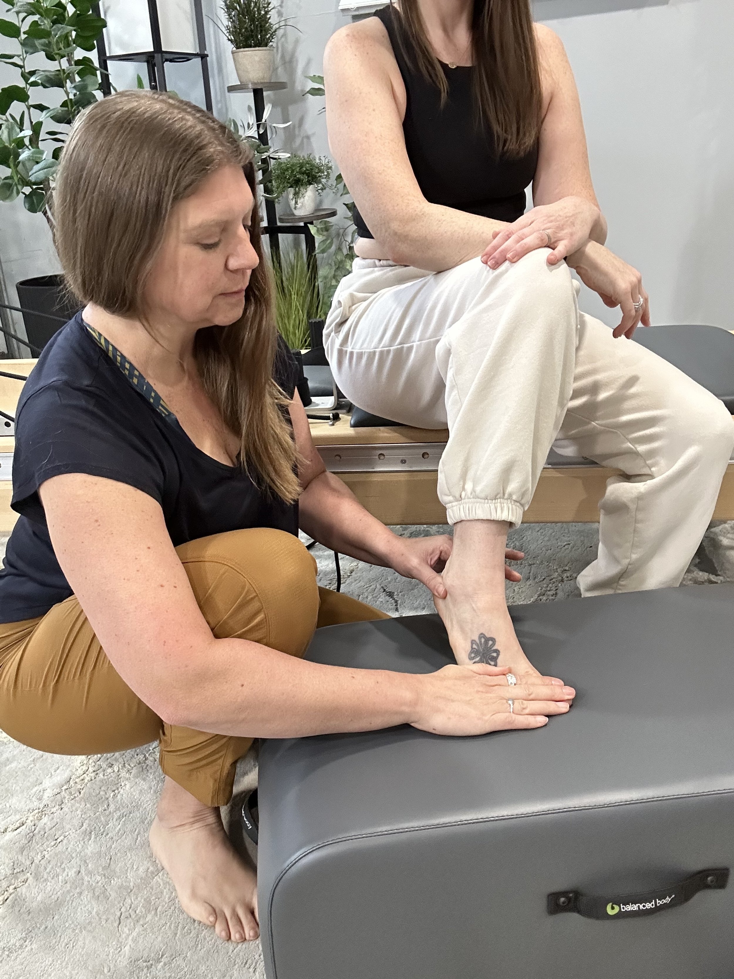 A woman is sitting on a therapy table with her leg extended. Another woman is kneeling beside her, holding her ankle and foot, possibly performing a physical therapy or massage. The setting appears to be a therapy or wellness clinic with plants and shelves in the background.