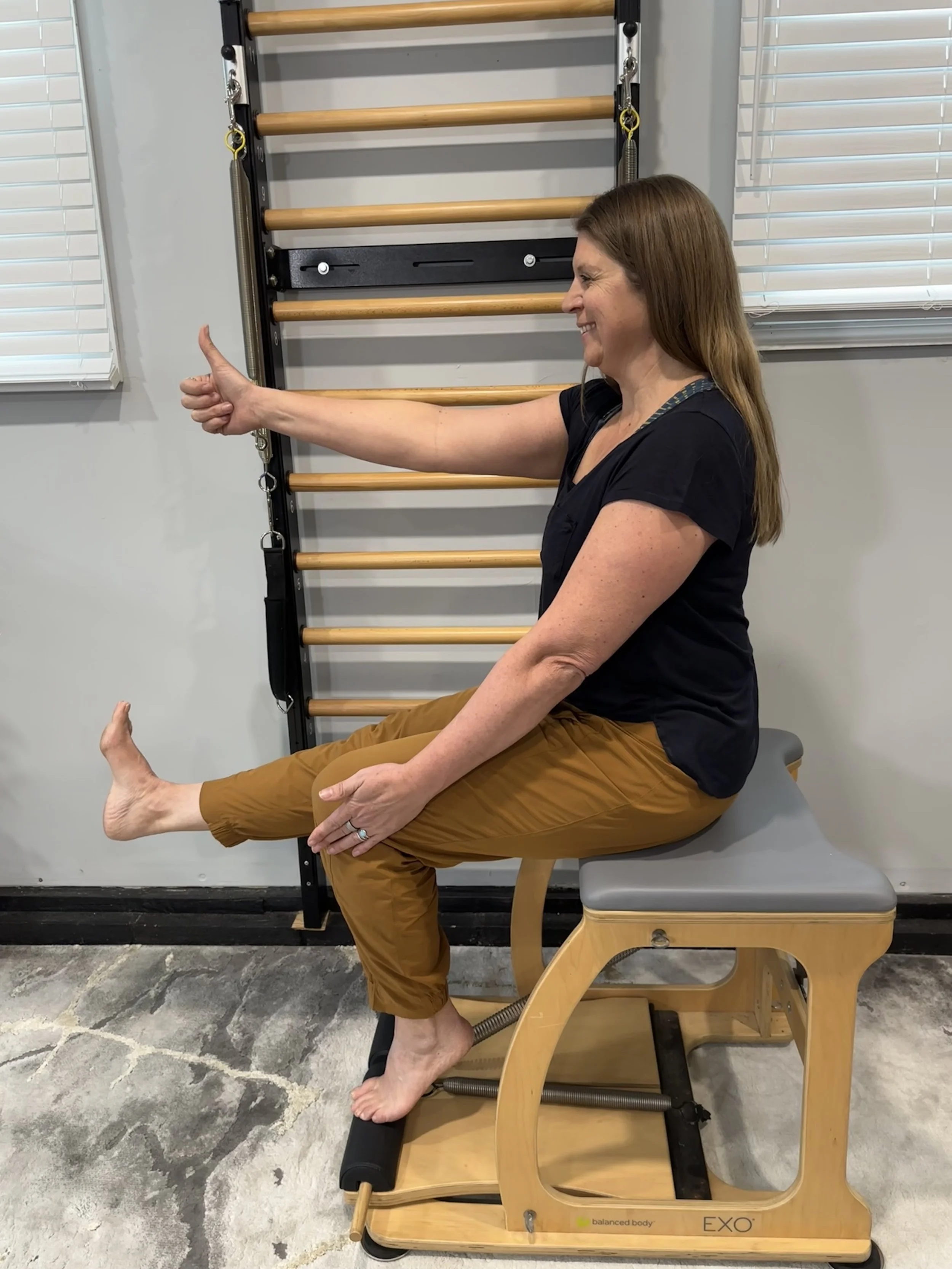 A woman sitting on a wooden fitness chair with electrical stimulation pads on her thigh, giving a thumbs up in front of a wall-mounted exercise ladder in a gym or therapy room.