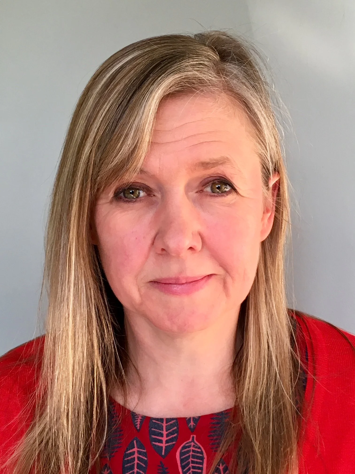 Portrait of a middle-aged woman with long blonde hair, wearing a red sweater and a blue patterned top, smiling slightly.