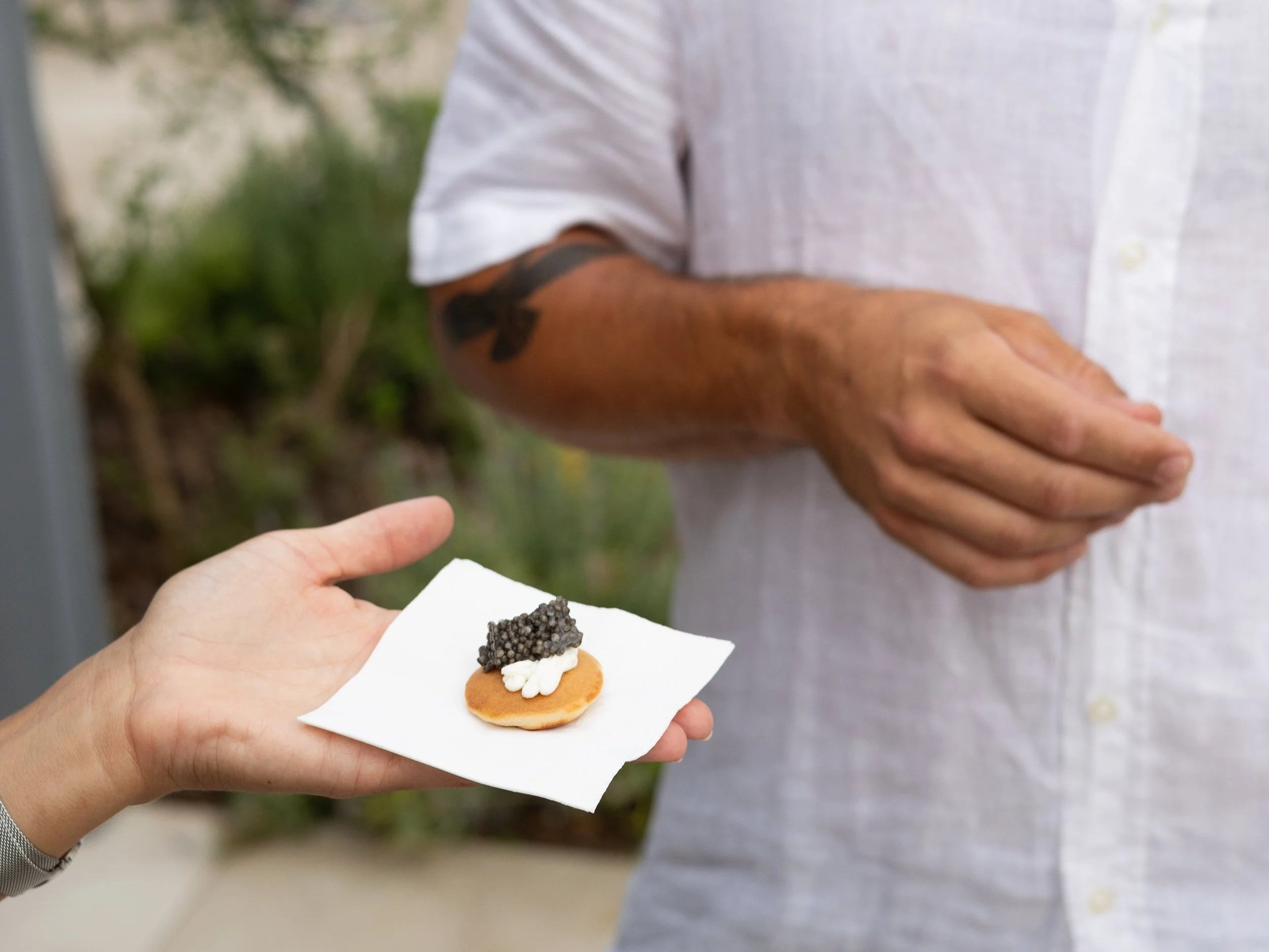 Person holding a small cracker with white cream topping and black caviar, while another person in white shirt points at it.