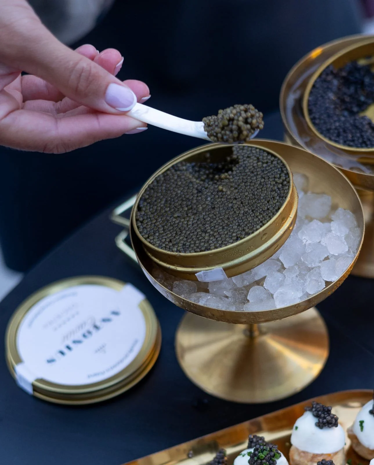 A hand holding a small spoon with caviar in front of open tins filled with black caviar on an ice bed, with some desserts topped with caviar in the foreground.