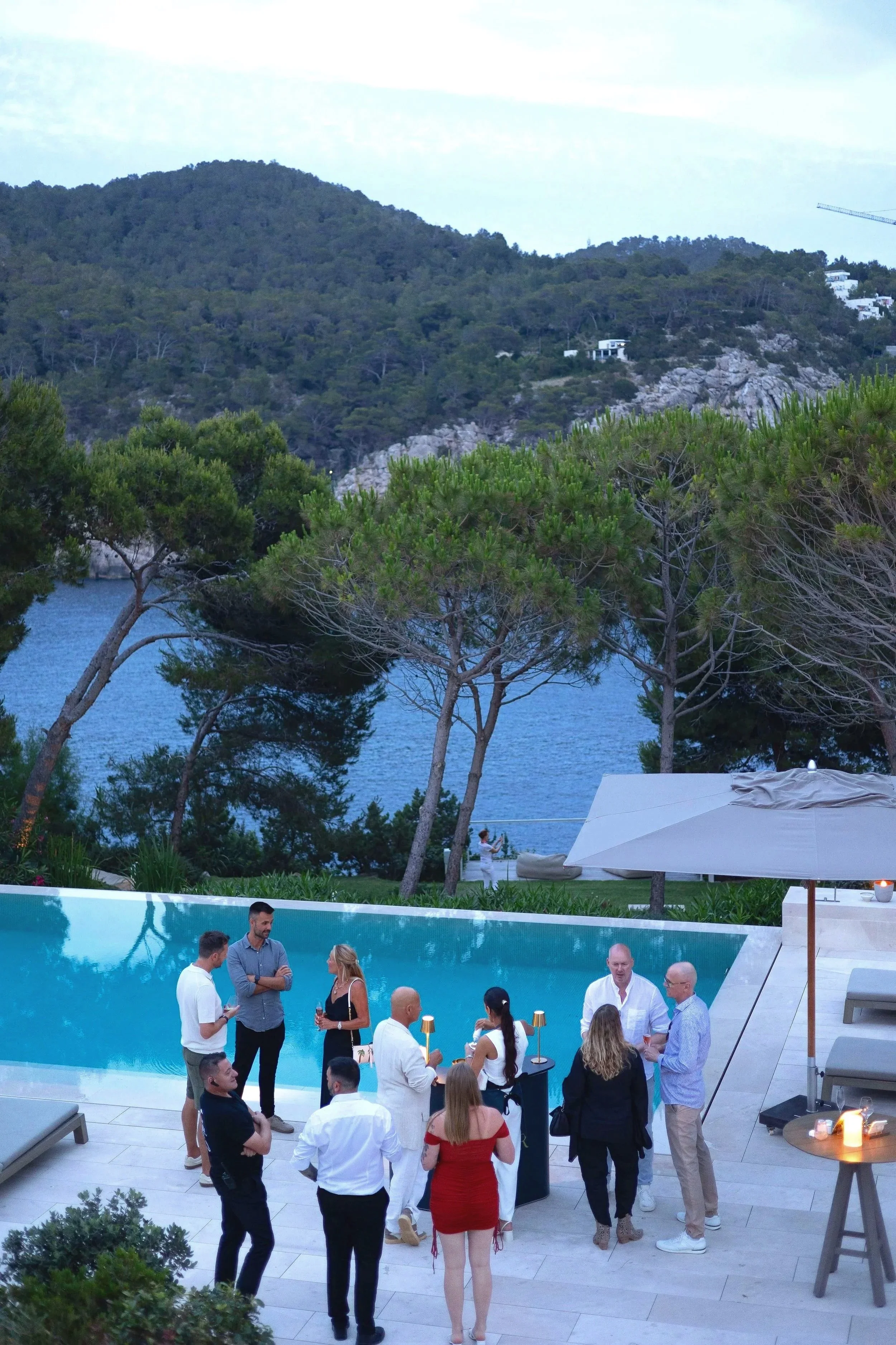 Group of people socializing by a swimming pool in villa Isla Feradura in Ibiza. A scenic outdoor setting with trees, hills, and water in the background at dusk.