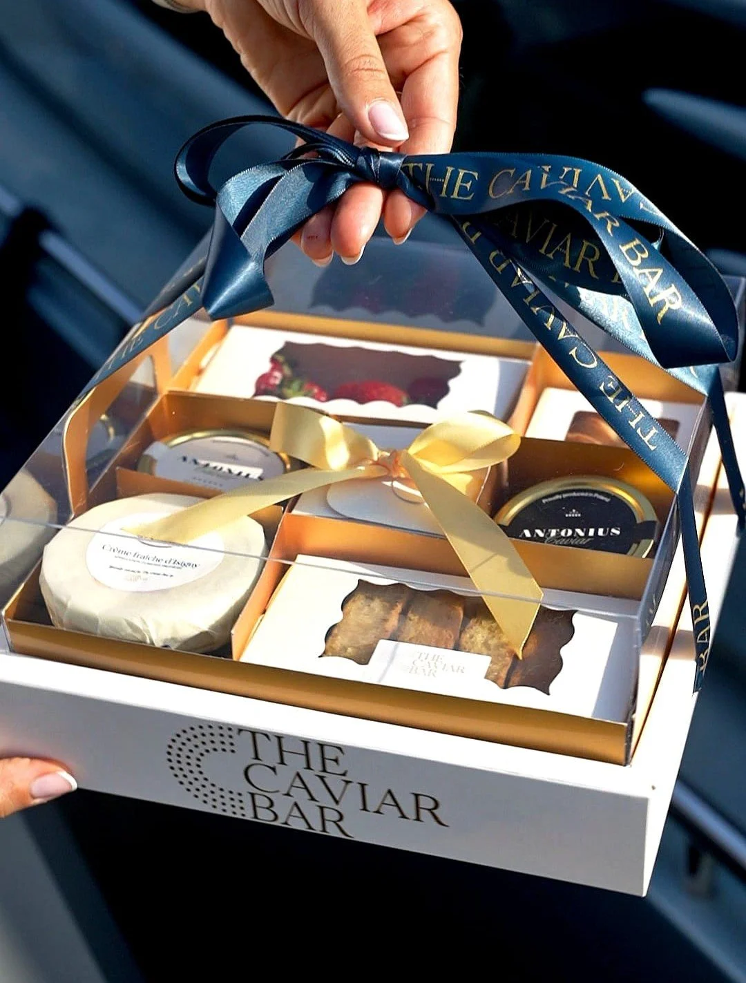 A hand holding a gift box labeled 'The Caviar Bar' with a dark blue ribbon, containing assorted gourmet items including caviar, smoked salmon, and baked goods, with decorative packaging and ribbons.