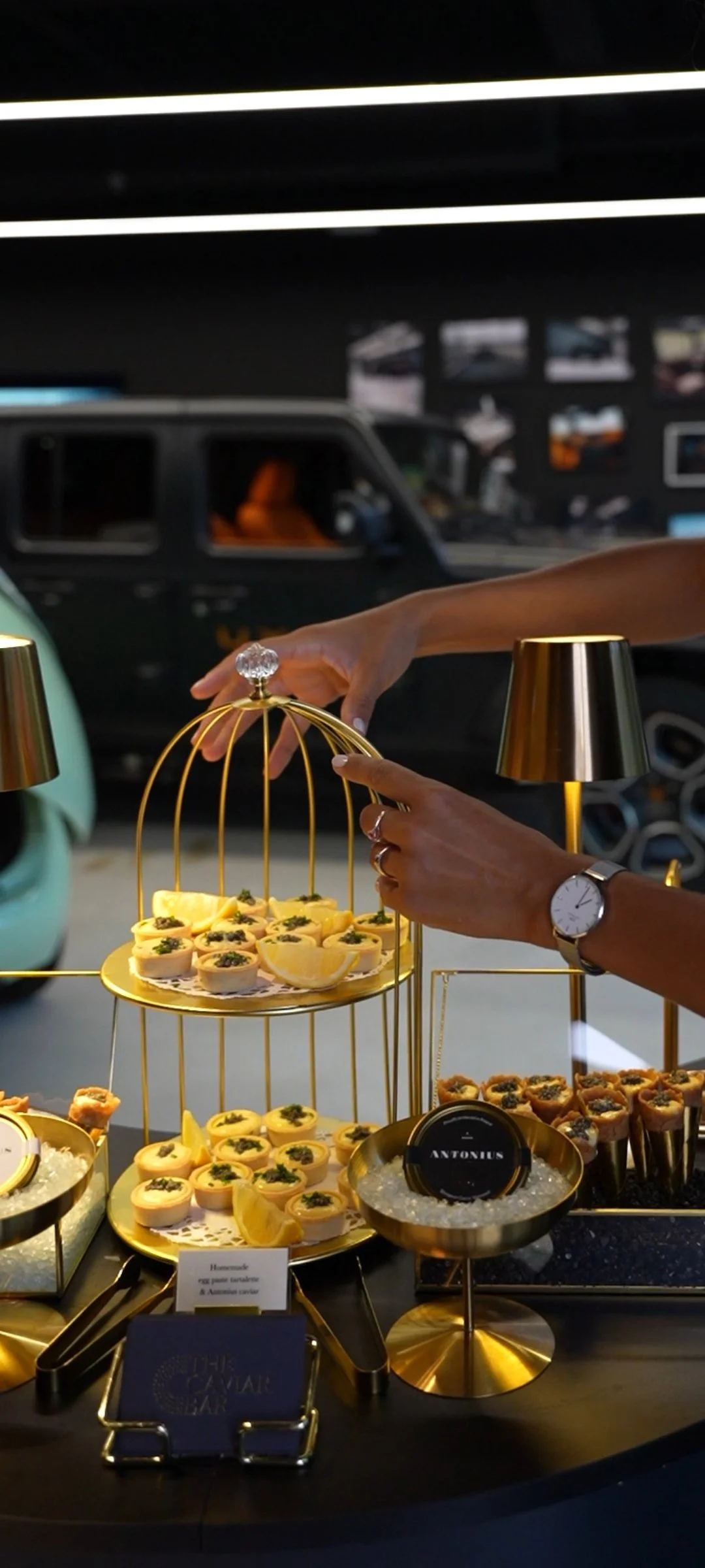 Hand arranging small appetizers on a gold tray at an event, with a blurred dark vehicle and photos in the background.