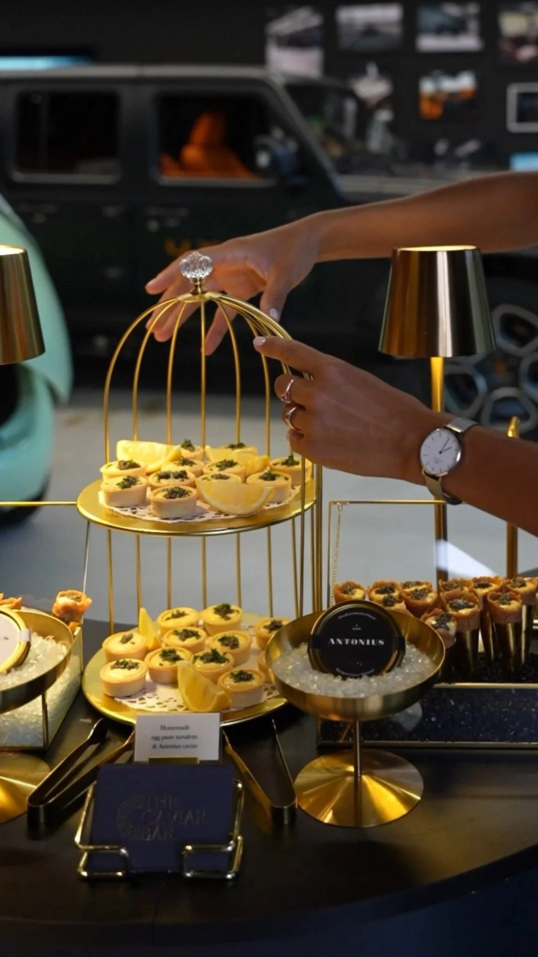 Elegant display of small appetizers and desserts on gold trays, set on a black table, with a person's hands reaching to pick one, and a background of cars and blurred interior.