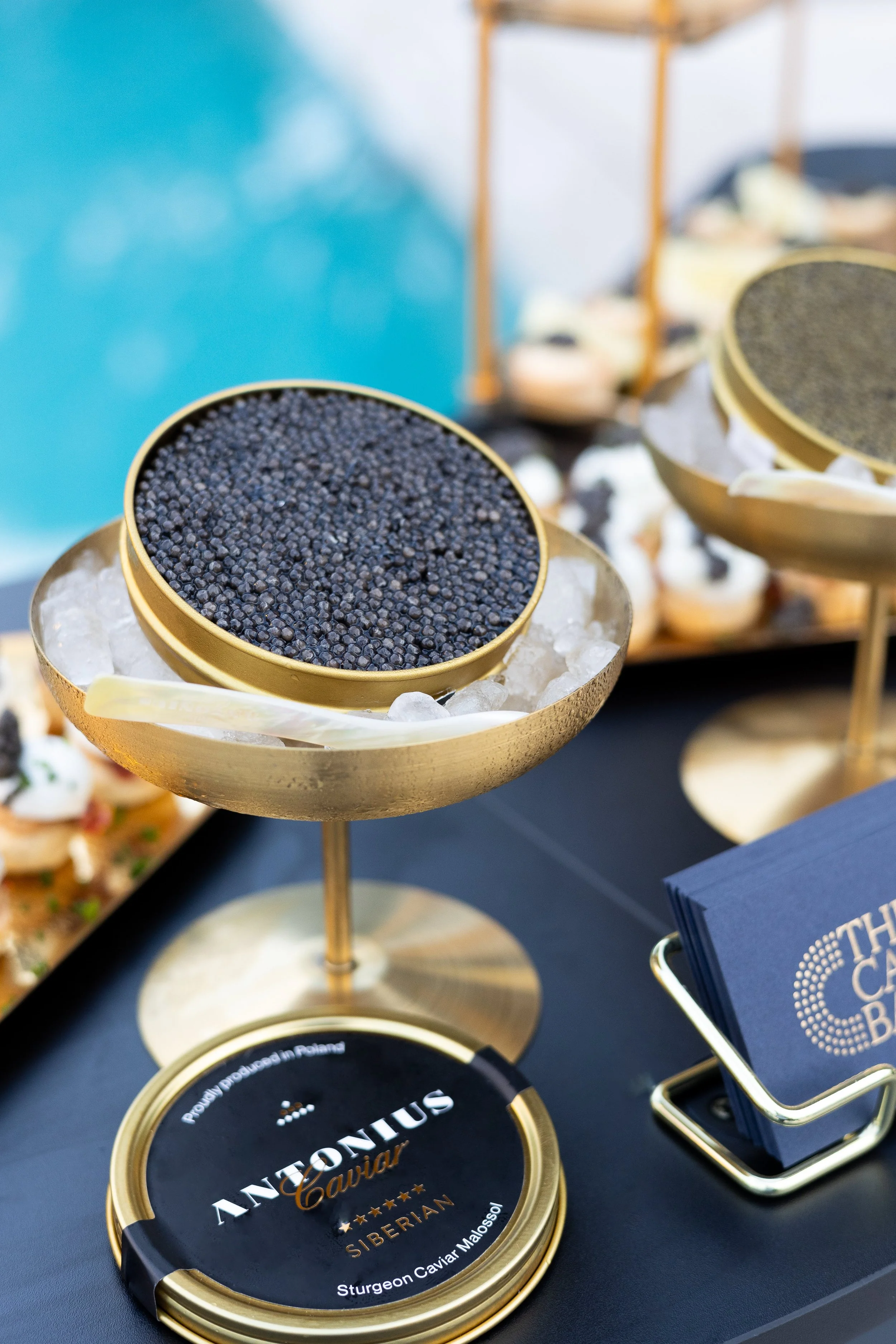 Close-up of a gold container of Antonis Caviar, Siberian type, on ice with other caviar containers and appetizers in the background.
