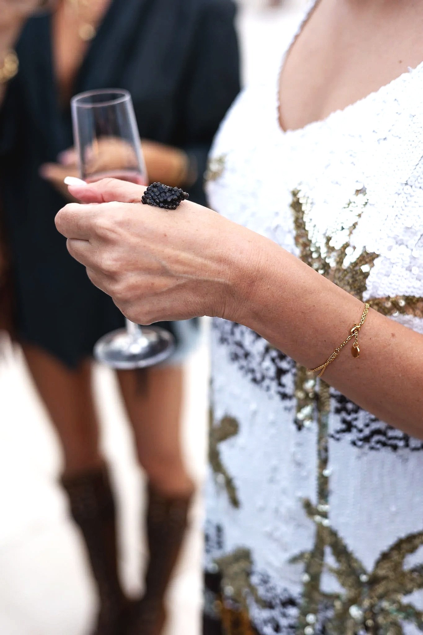 Close-up of woman's hand with a large black ring, holding a champagne flute and caviar bump at a social event with people in the background. Isla Sa Feradura Ibiza.