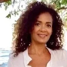 A woman with curly hair smiling outdoors against a backdrop of trees and water.