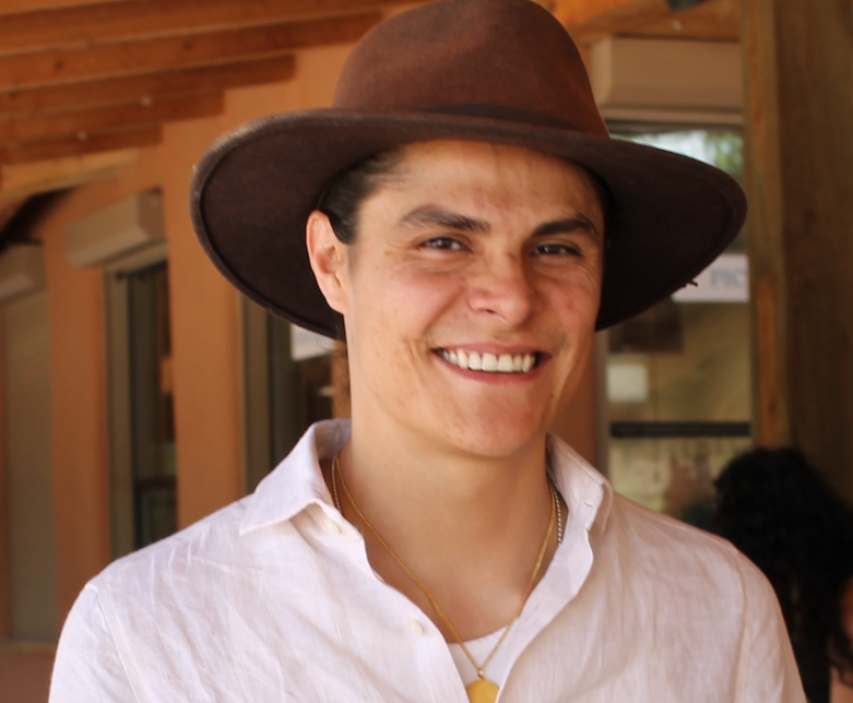 A young man wearing a large brown hat and white shirt, smiling outdoors with a wooden structure in the background.