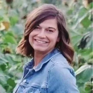 A woman with shoulder-length brown hair smiling outdoors, wearing a denim jacket with greenery in the background.