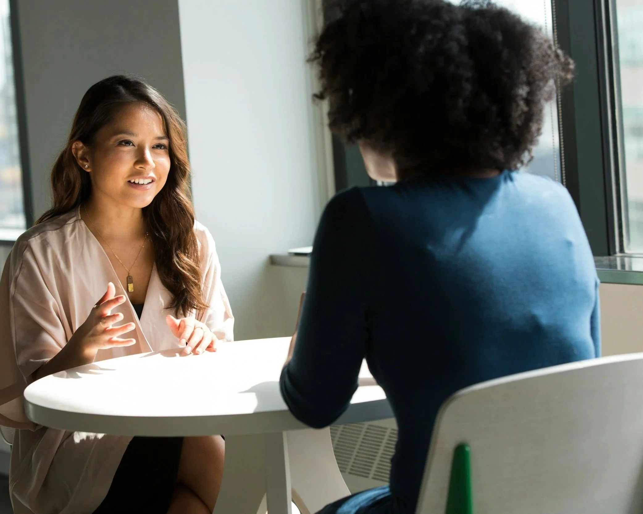 Two women sitting across from each other at a small, white table in a bright room, engaging in a conversation. One woman, with light skin and long, wavy brown hair, is speaking, while the other woman, with darker skin and curly hair, is listening. They appear to be in a professional or casual meeting near a window with natural light.