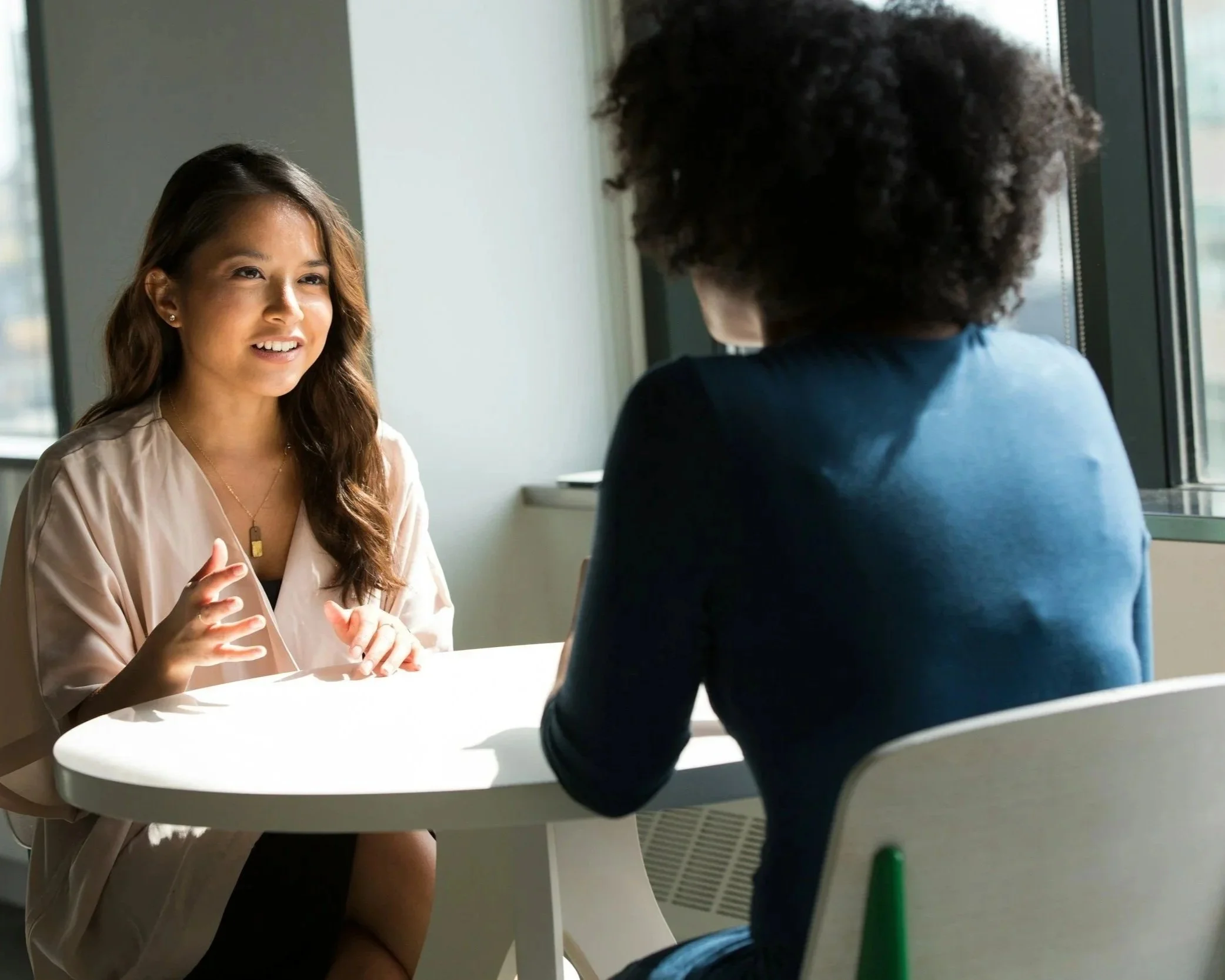 Two women having a conversation at a table near a window, with sunlight illuminating their faces.