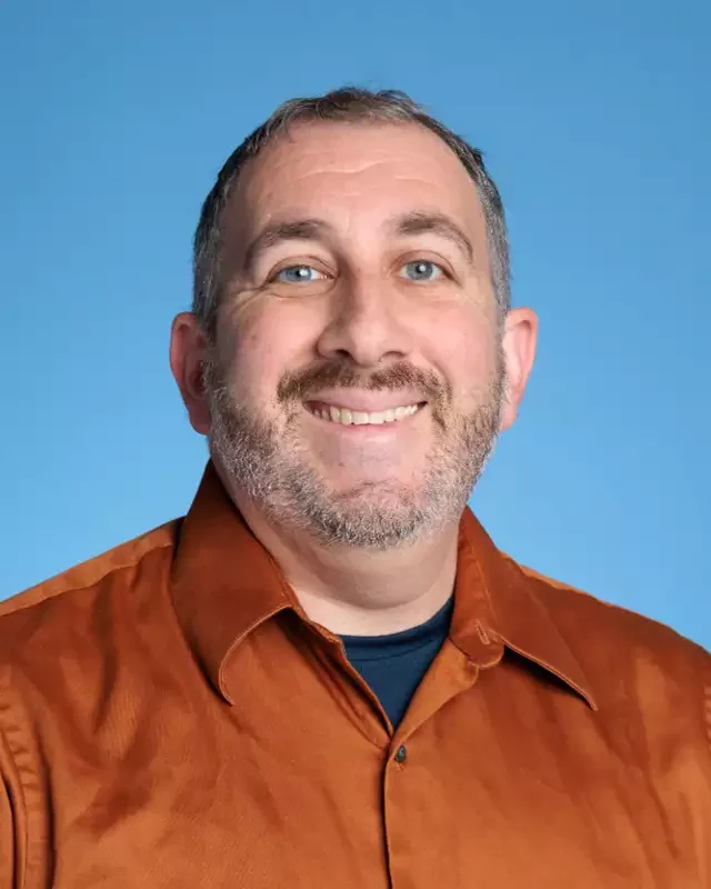 A smiling man with short gray hair and a beard, wearing a brown button-up shirt over a black t-shirt, standing against a blue background.