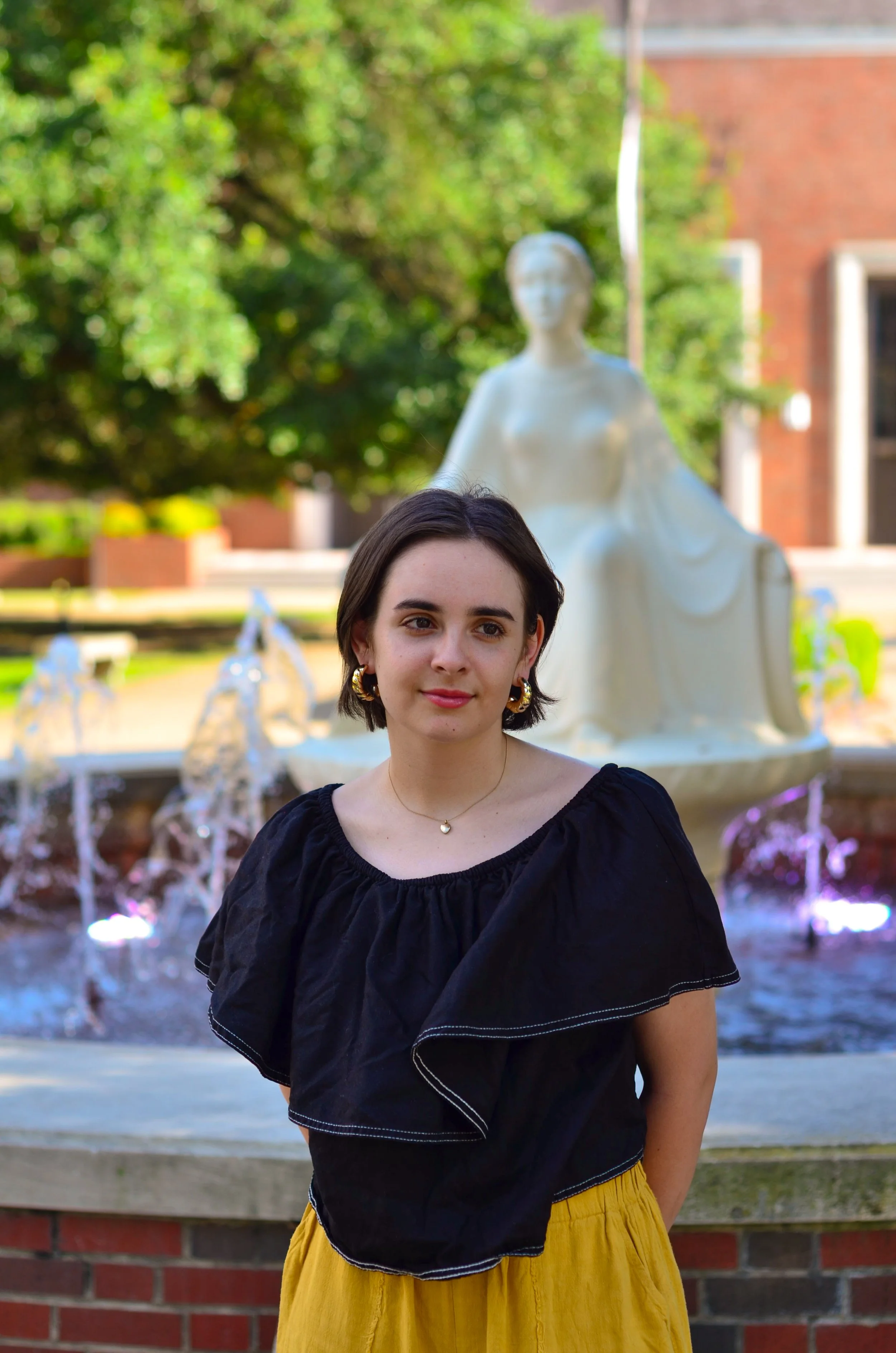 A young woman with short dark hair, wearing a black ruffled top and yellow skirt, standing in front of a decorative water fountain with a white statue of a seated woman in a public garden.