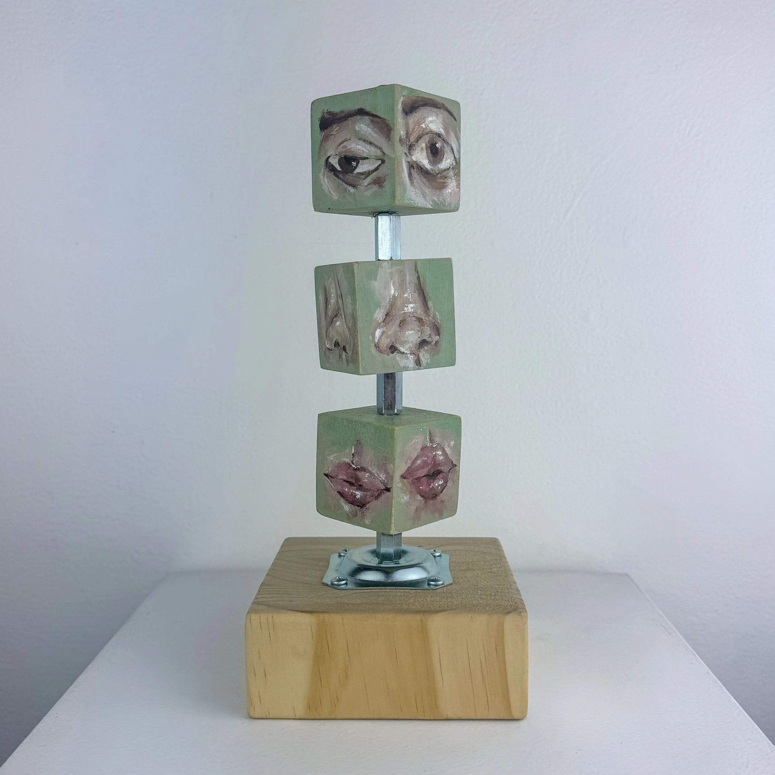 A sculpture with four painted wooden cubes mounted on a metal rod, with faces of a person on the cubes and lips on the bottom cube, positioned on a wooden block base.