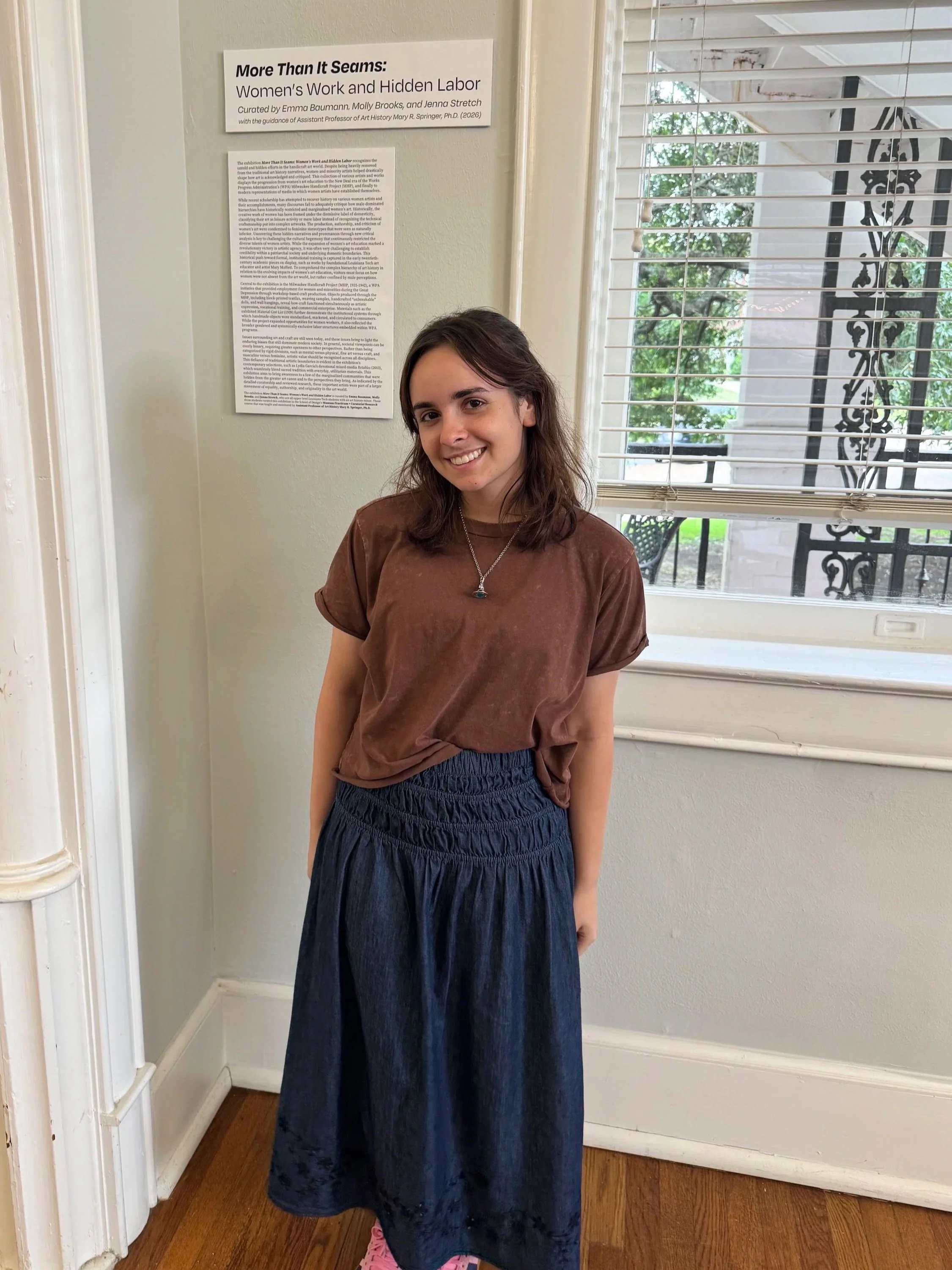 Emma Baumann standing beside the exhibition statement for More Than It Seems: Women's Work and Hidden Labor, smiling toward the camera in a gallery with large windows.