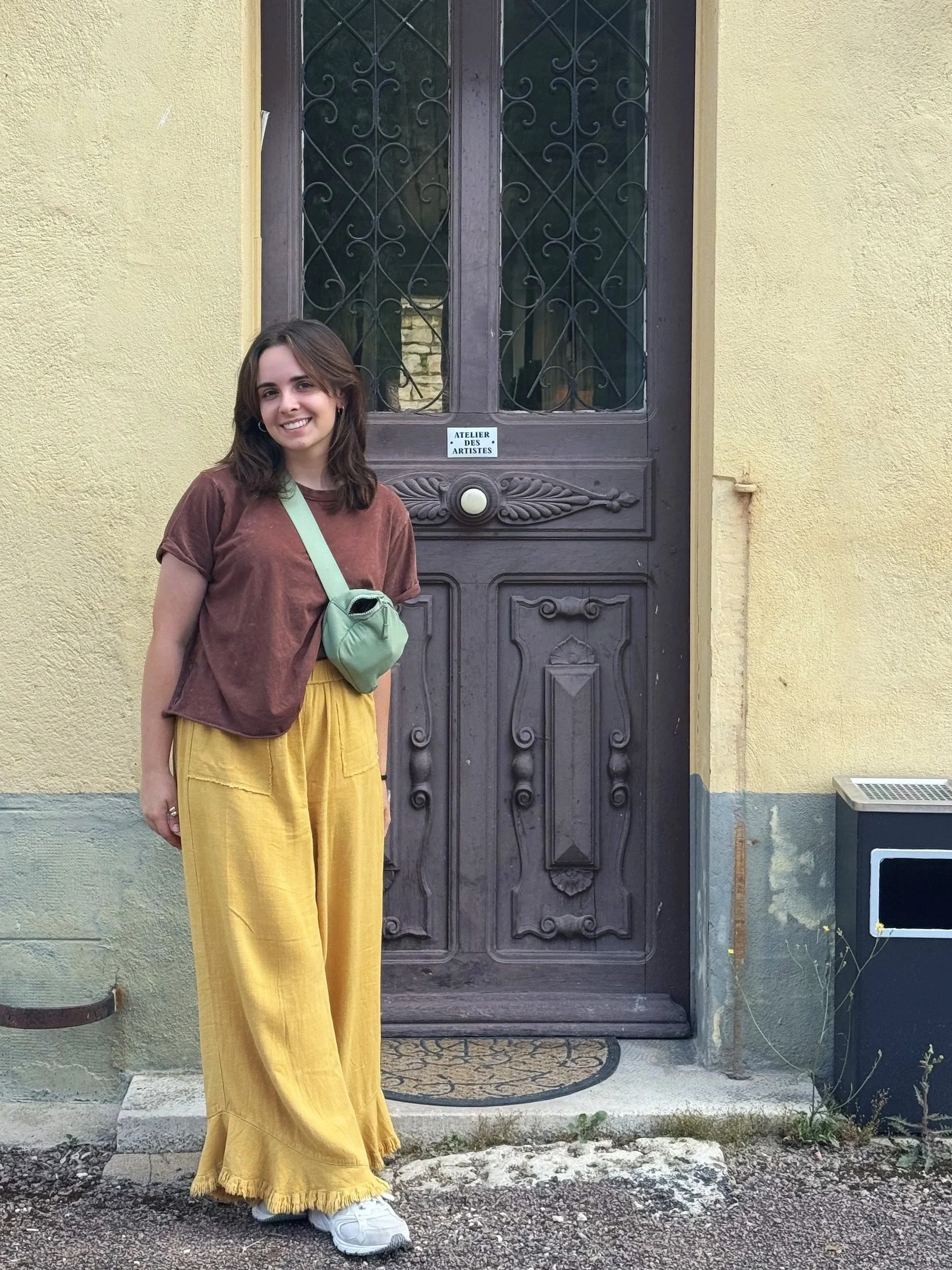 Emma Baumann standing in front of a tall French door with decorative wrought-iron glass panels set into a pale yellow stucco wall. She wears a brown shirt, loose mustard-yellow pants, and a green crossbody bag, smiling toward the camera.