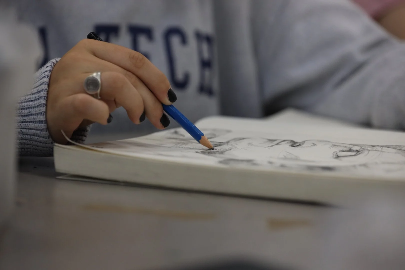 Close-up of a person drawing sketches in a sketchbook with a blue pencil, wearing a gray sweatshirt with visible black painted nails and a ring on their finger.
