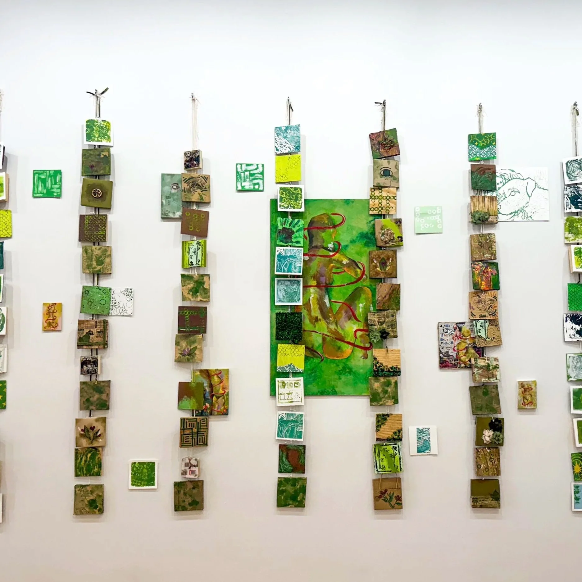 An art display with multiple small, square artworks arranged in vertical columns on a white wall, with a central larger painting featuring abstract green shapes and red lines.