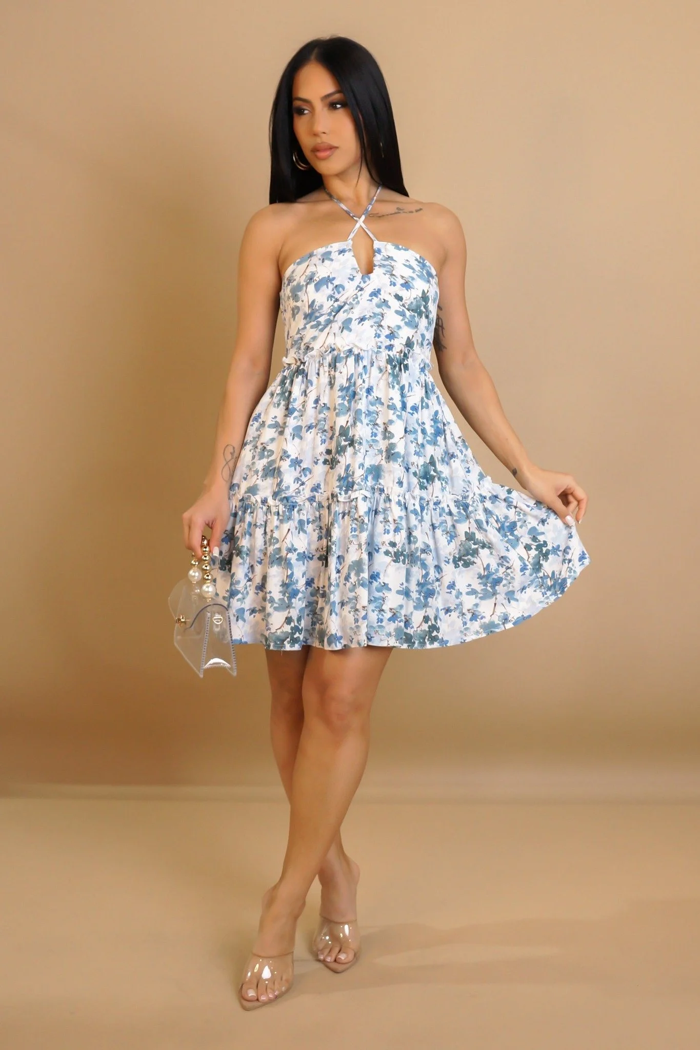A woman with long black hair wearing a white and blue floral dress, clear heeled shoes, and holding a handbag with pearl accents, standing in front of a beige background.
