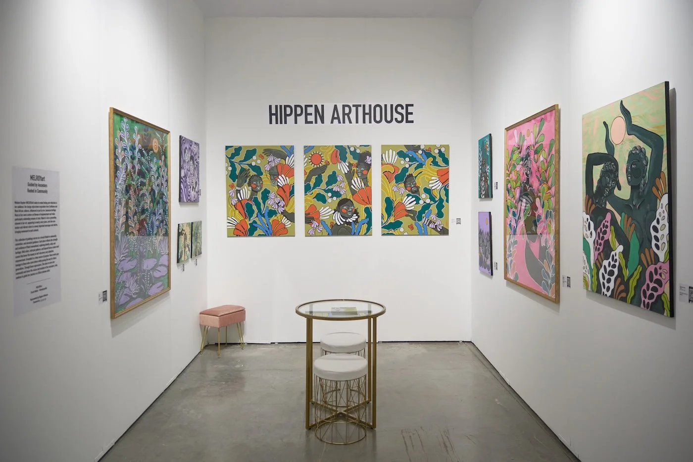 Art gallery with colorful, modern paintings featuring abstract and stylized floral and human figures, white walls, small table with two stools, and the sign 'HIPPEN ARTHOUSE' on the wall.