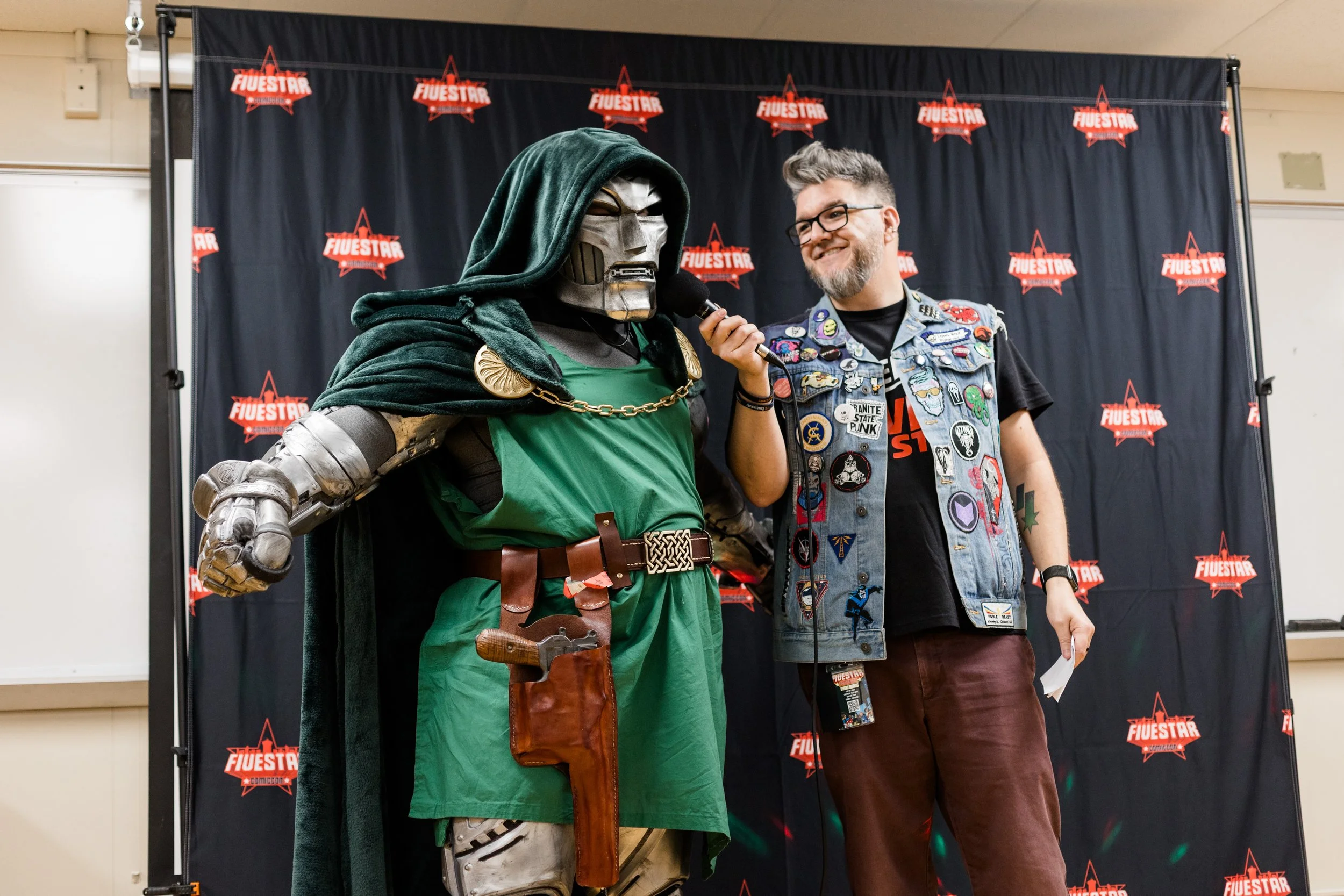 A person in a robot costume dressed as a sci-fi character being interviewed at a comic convention, with a man holding a microphone. The background has a black banner with red and white "Fuestar" logos.