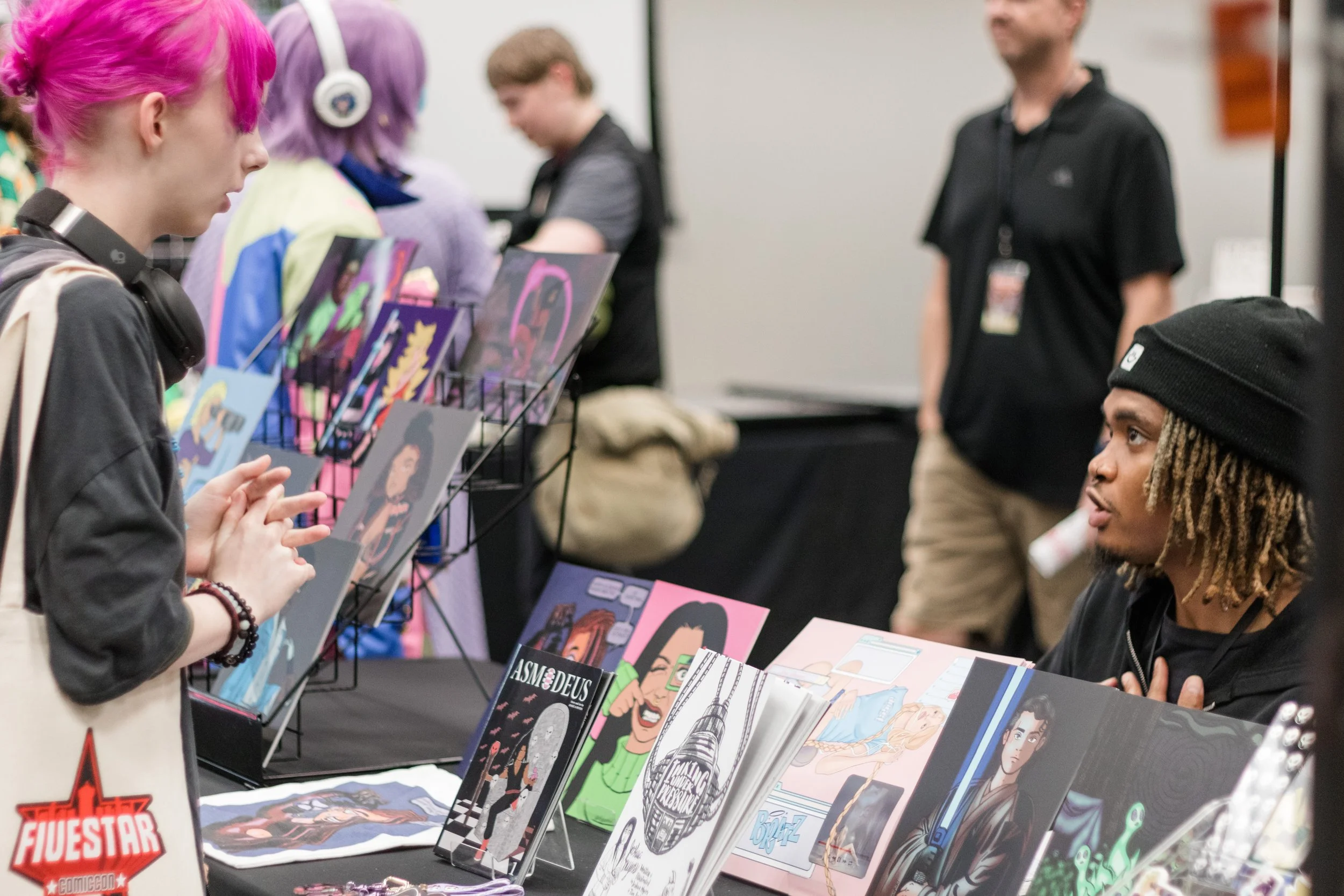 A young woman with pink hair looking at artwork and comic book prints at a convention booth, with colorful artwork displayed on the table and a seller speaking to her.