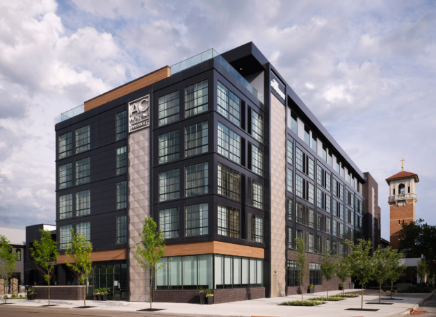 Modern multi-story office building with large glass windows and a sign that reads 'AC' on the upper corner, with a church tower visible in the background, under a partly cloudy sky.