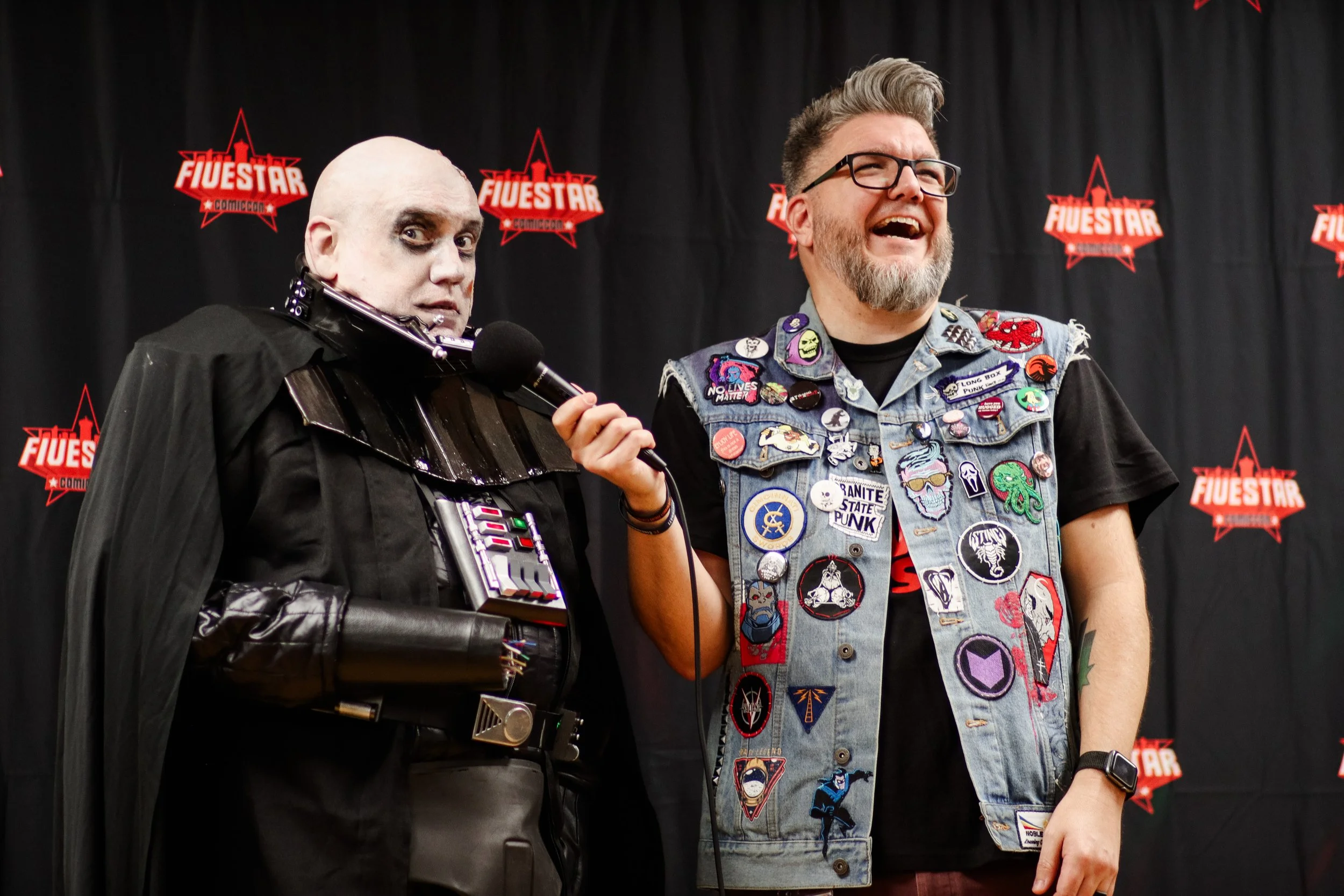 Two men standing in front of a black backdrop with red 'Fuestar Comiccon' logos. The man on the left dressed as a character with makeup, a black cape, and a costume with a device on the chest, holding a microphone. The man on the right is smiling, we