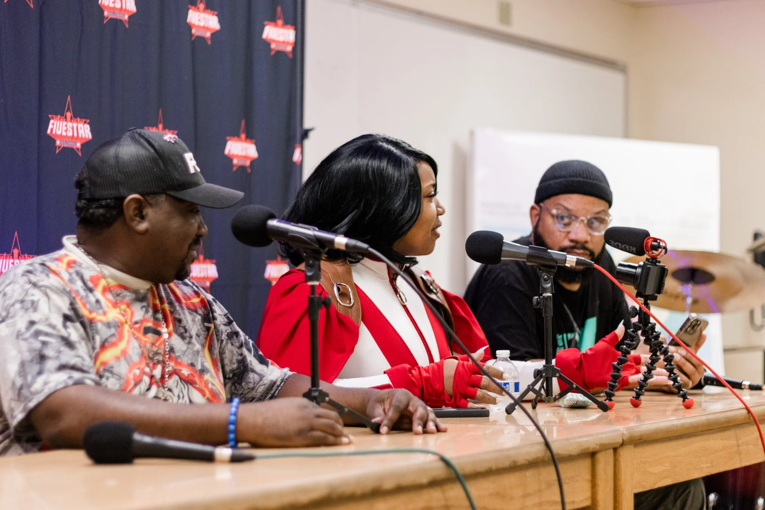 Three people sitting at a table, participating in a panel discussion or interview. All are equipped with microphones. The background features a navy blue curtain with red and white logos.