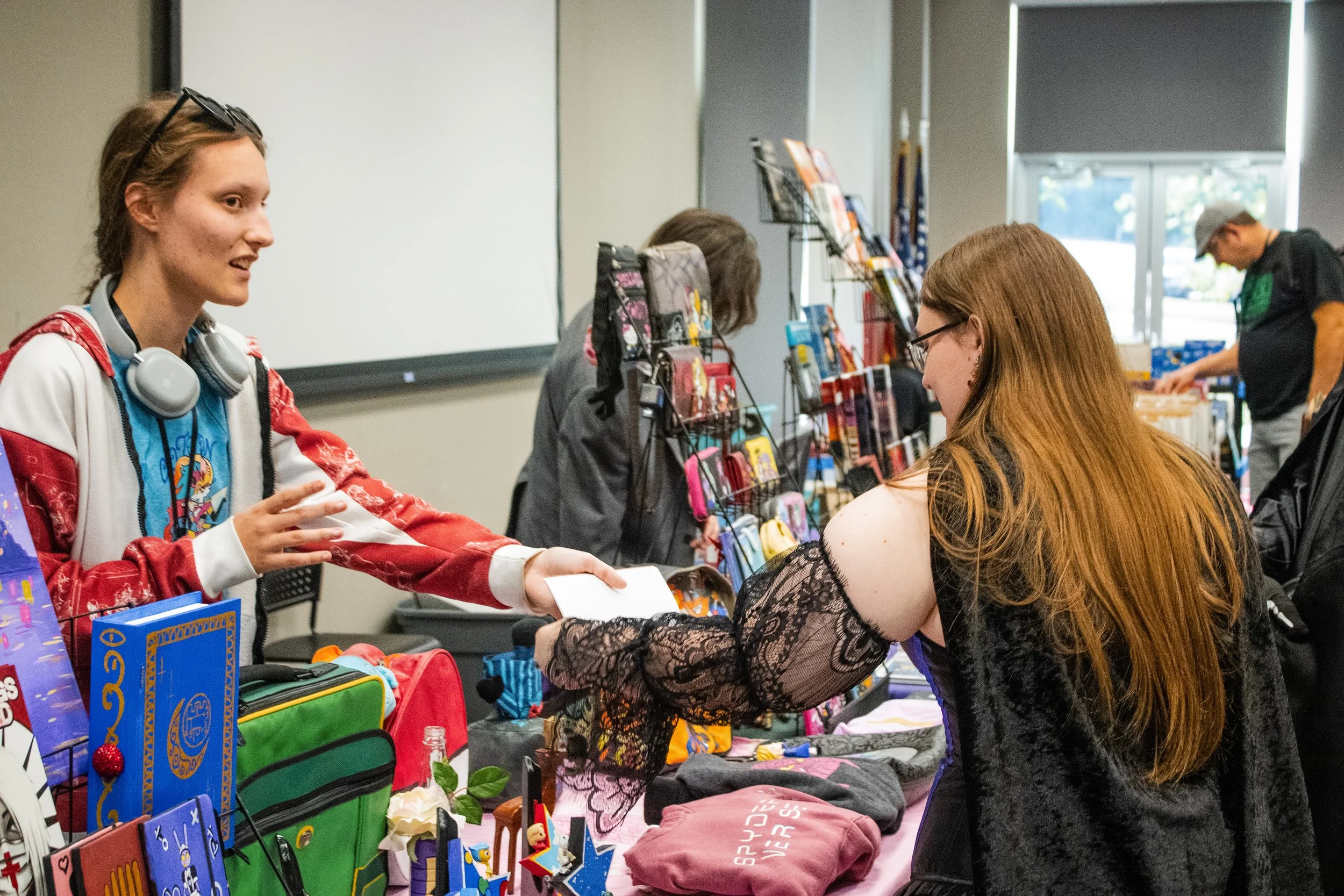 A woman with long red hair and glasses is shopping at a table of small items, including clothing and accessories, while a sales associate with short hair, glasses on her head, and headphones around her neck exchanges conversation or a purchase.