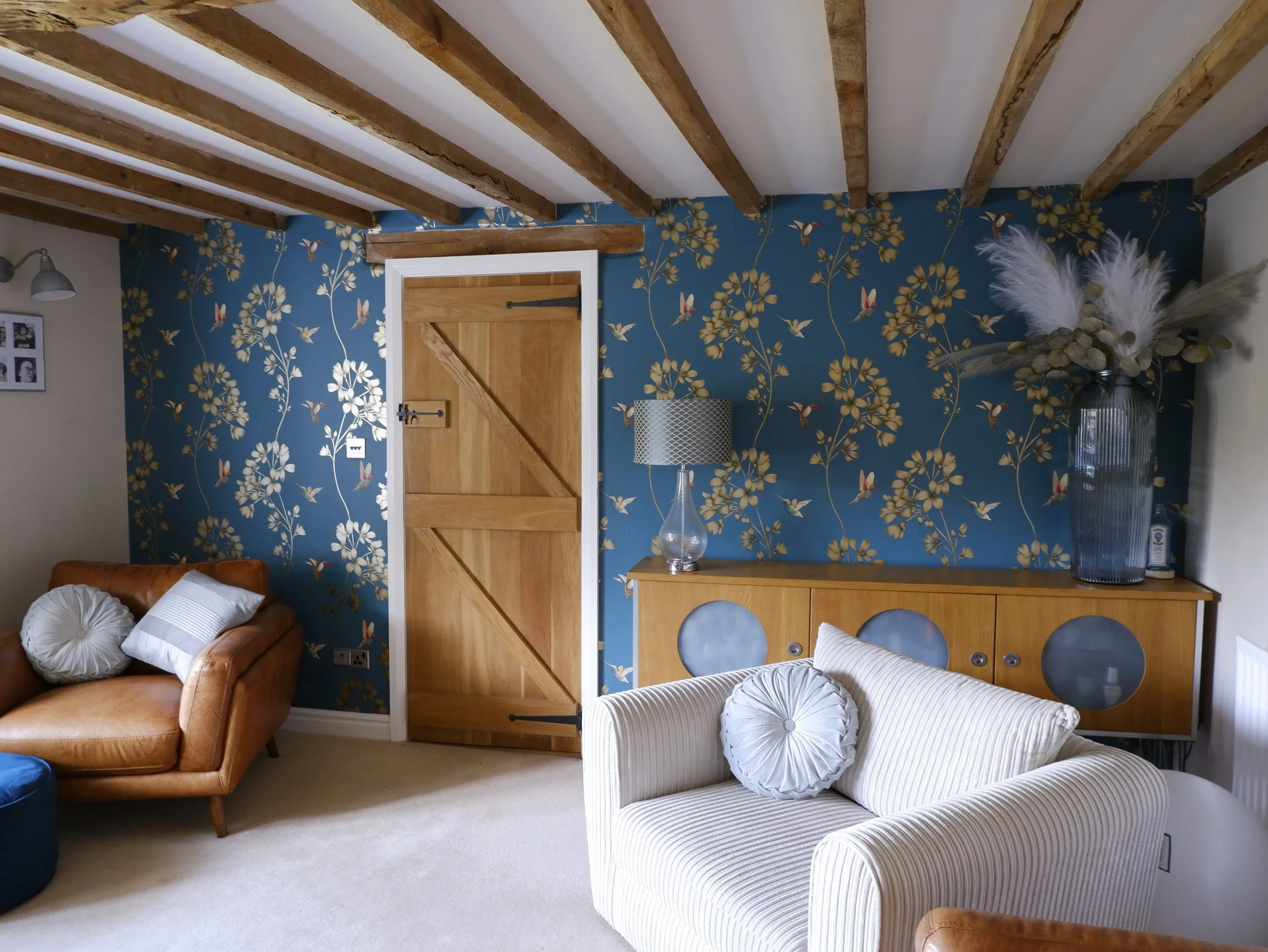 Living room with a blue floral and bird wallpapered wall, wooden beams on the ceiling, a wooden door, a tan leather sofa with pillows, a white striped armchair with a round pillow, a wooden sideboard with a glass lamp and a large vase with dried flowers.