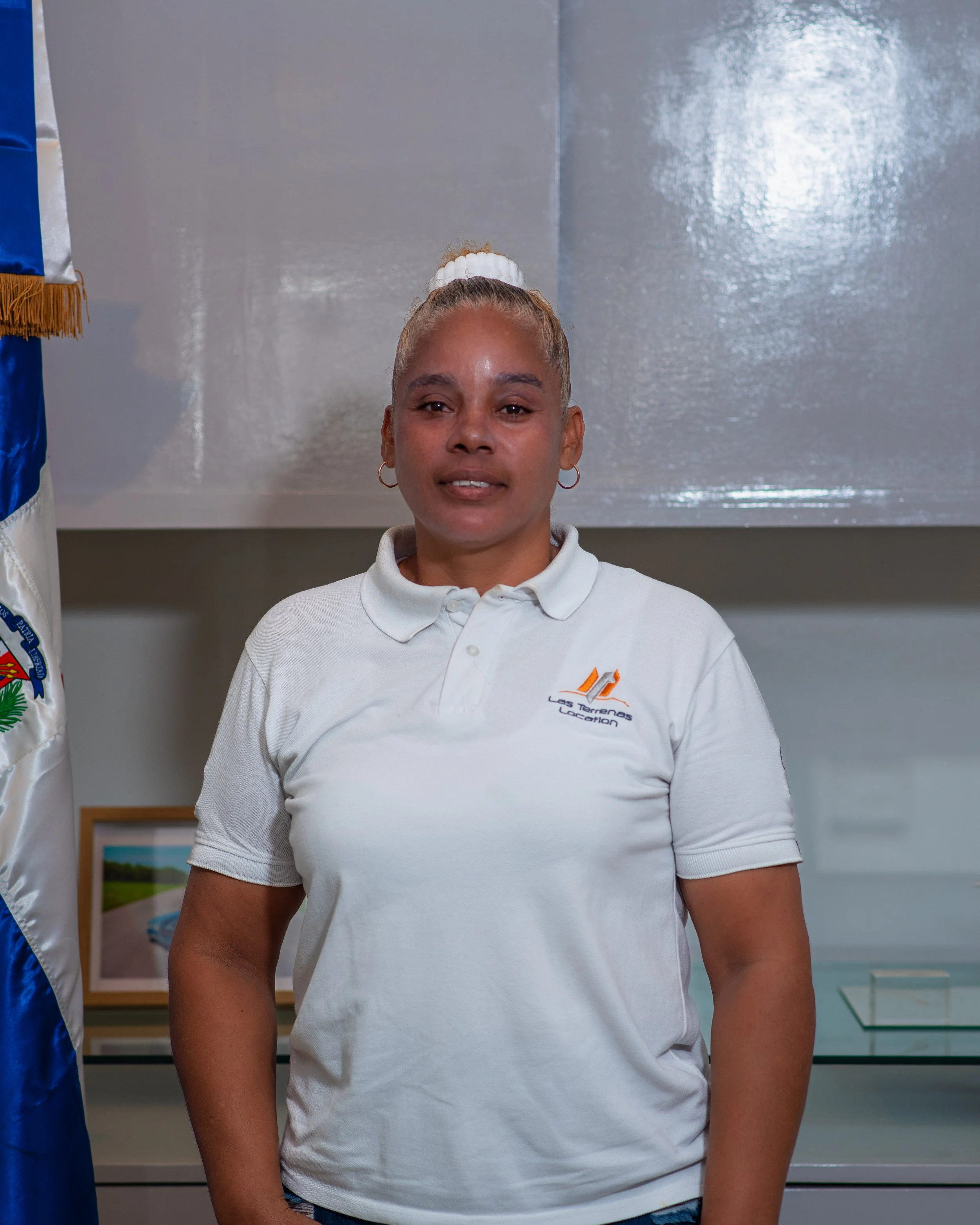 Mujer de pelo rubio atado con coleta, usando camisa blanca con logo en el pecho, de pie en interior con fondo gris y bandera a la izquierda.